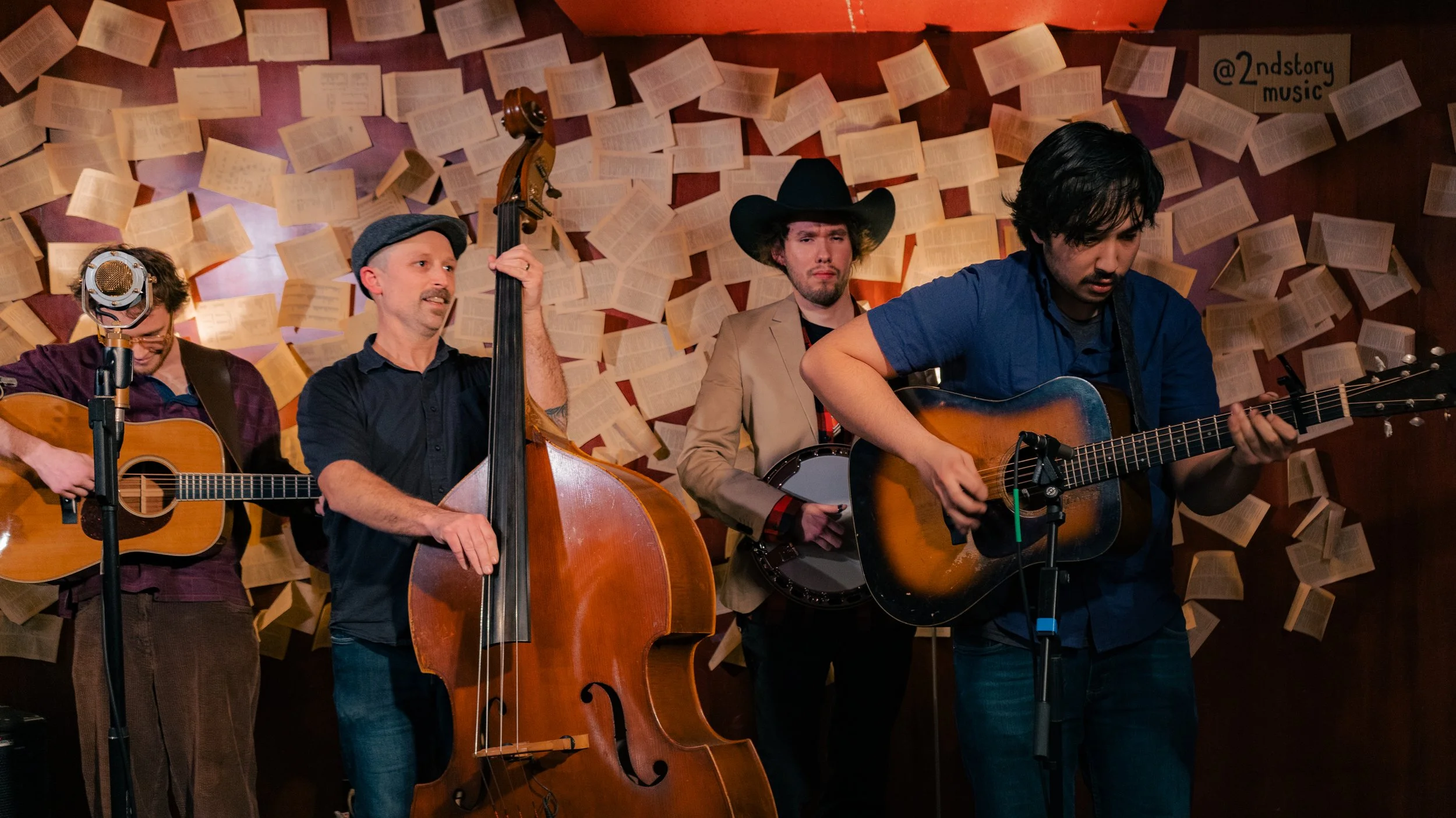 Four musicians performing with guitars, a double bass, and a banjo in front of a wall decorated with open books and a sign that reads '@2ndstory music'.