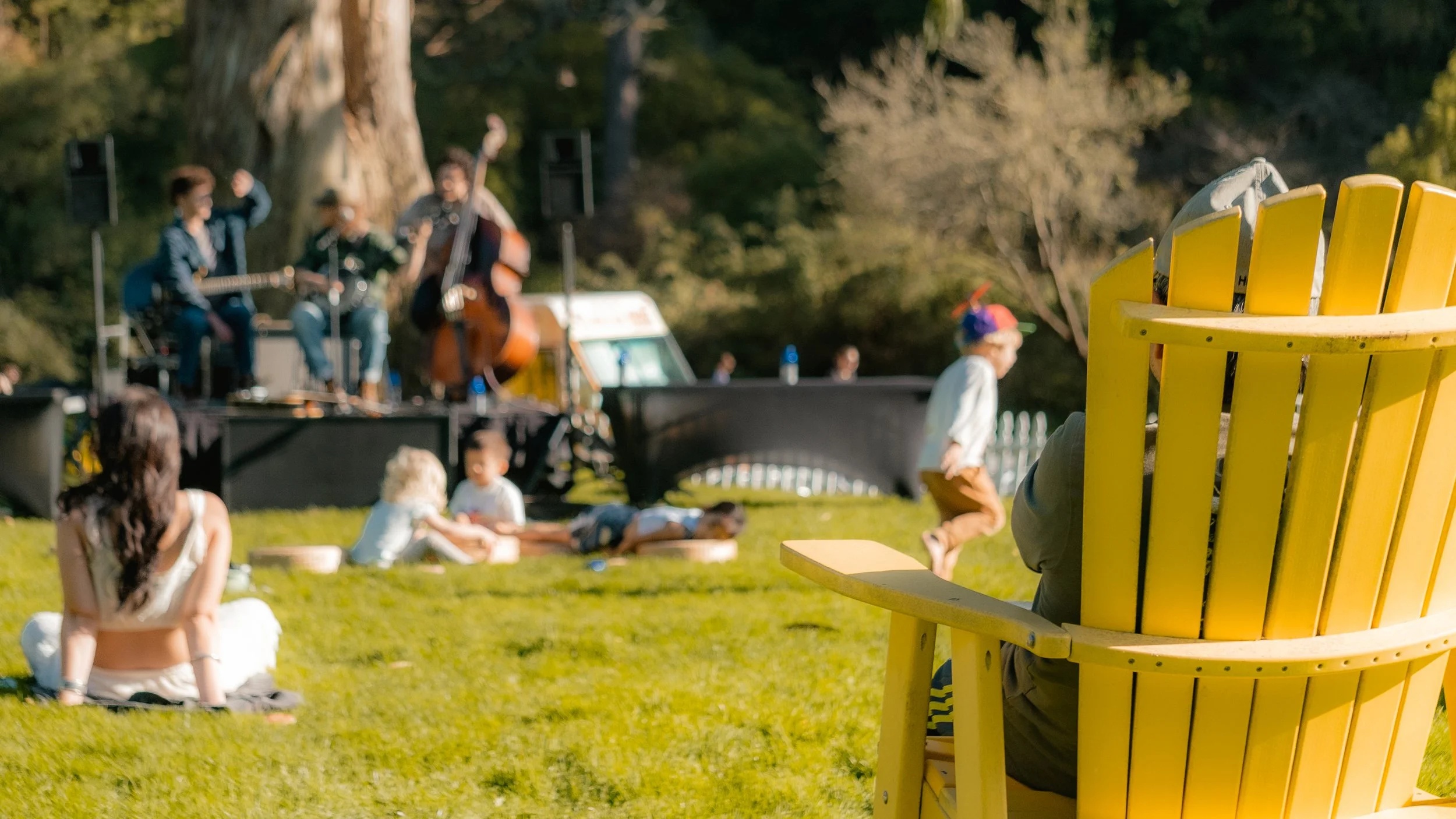 Person sitting on a large yellow Adirondack chair, facing an outdoor stage with a band performing and children playing on the grass in a park.