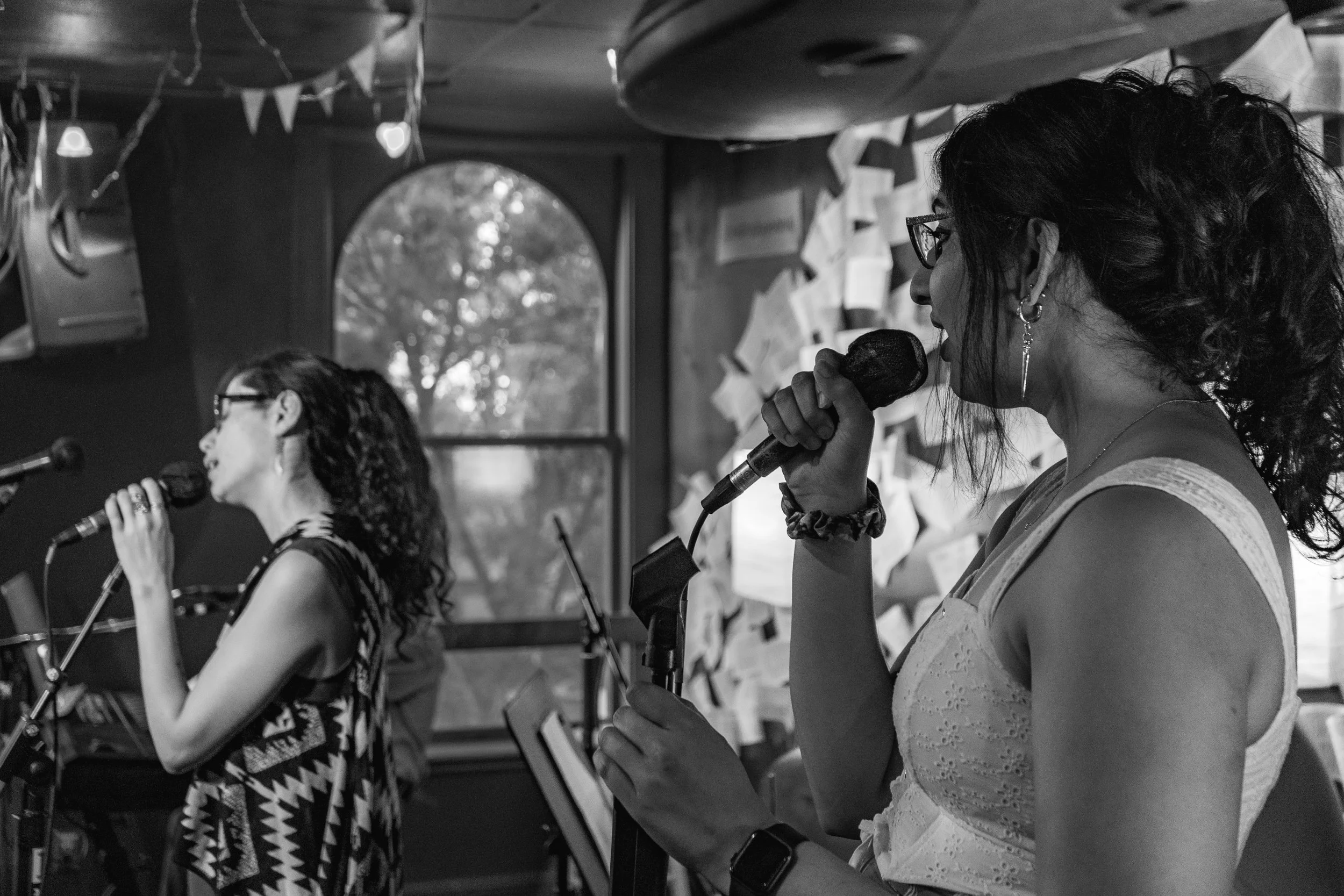 Two women singing into microphones in a room decorated with bunting and papers on the wall; a window with trees visible outside.