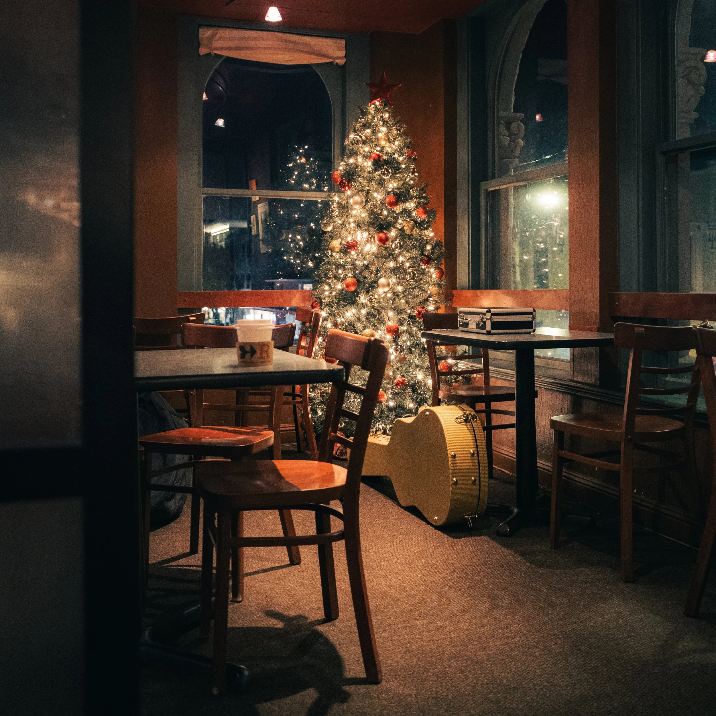 A decorated Christmas tree with lights and ornaments inside a cozy restaurant or cafe, with tables and chairs nearby and a large window reflecting the tree at night.