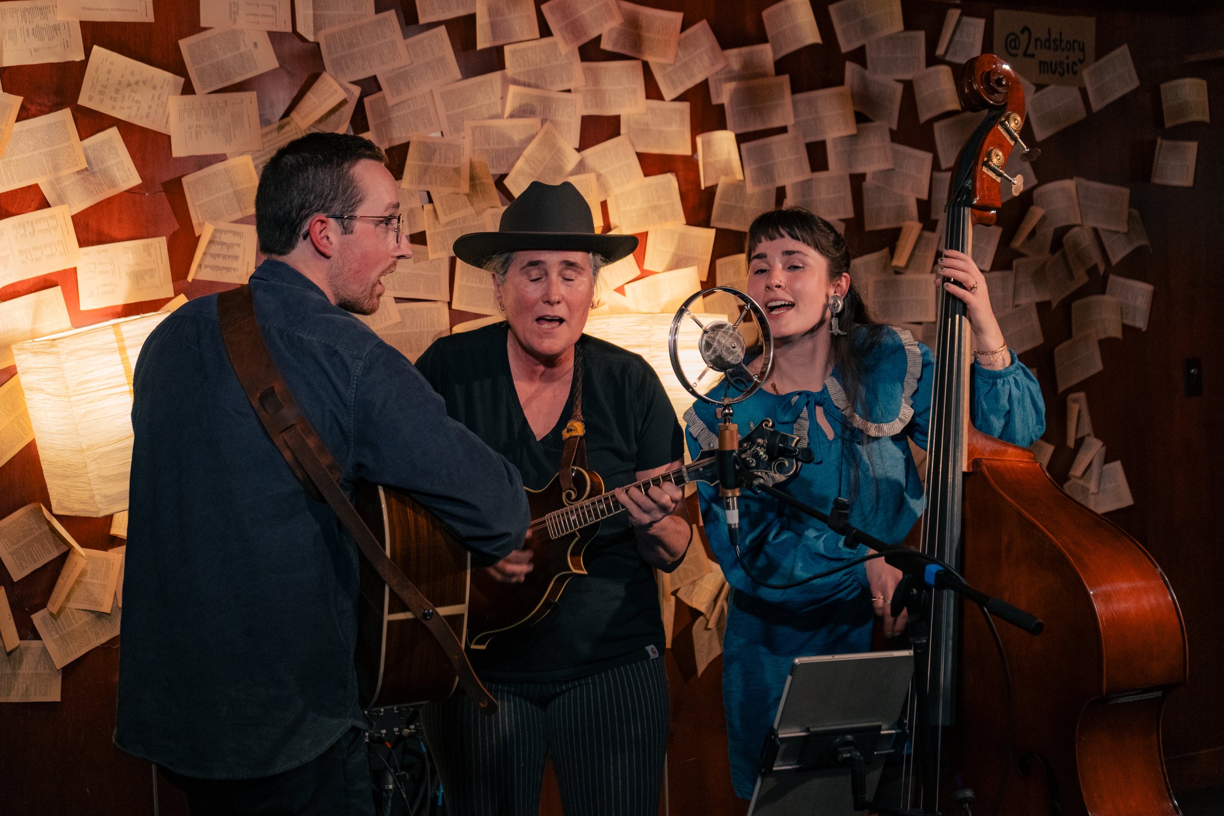 Three musicians singing and playing instruments in a cozy room with open books on the wall. The woman in the middle is playing a guitar, the woman on the right is playing a double bass, and the man on the left is playing a guitar.