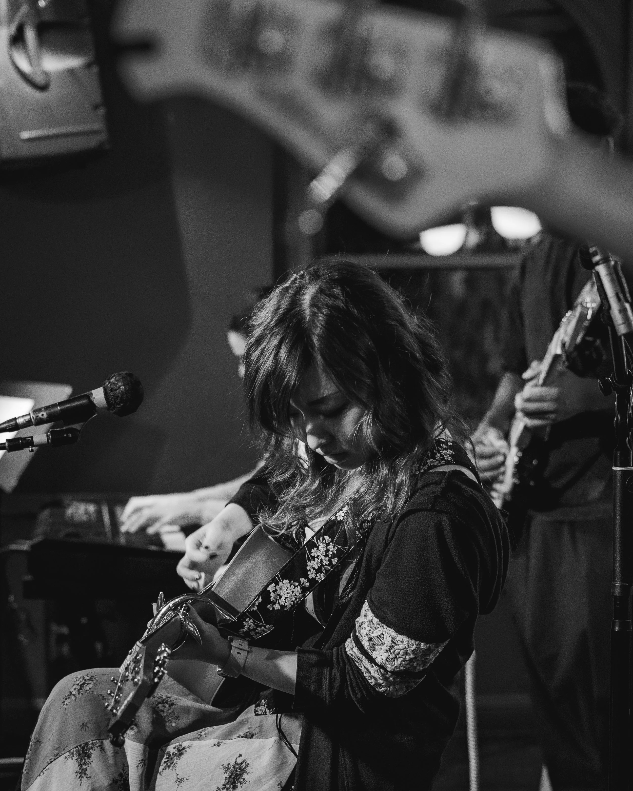 A woman with wavy hair playing an acoustic guitar during a performance, with other musicians in the background, in a dimly lit setting.