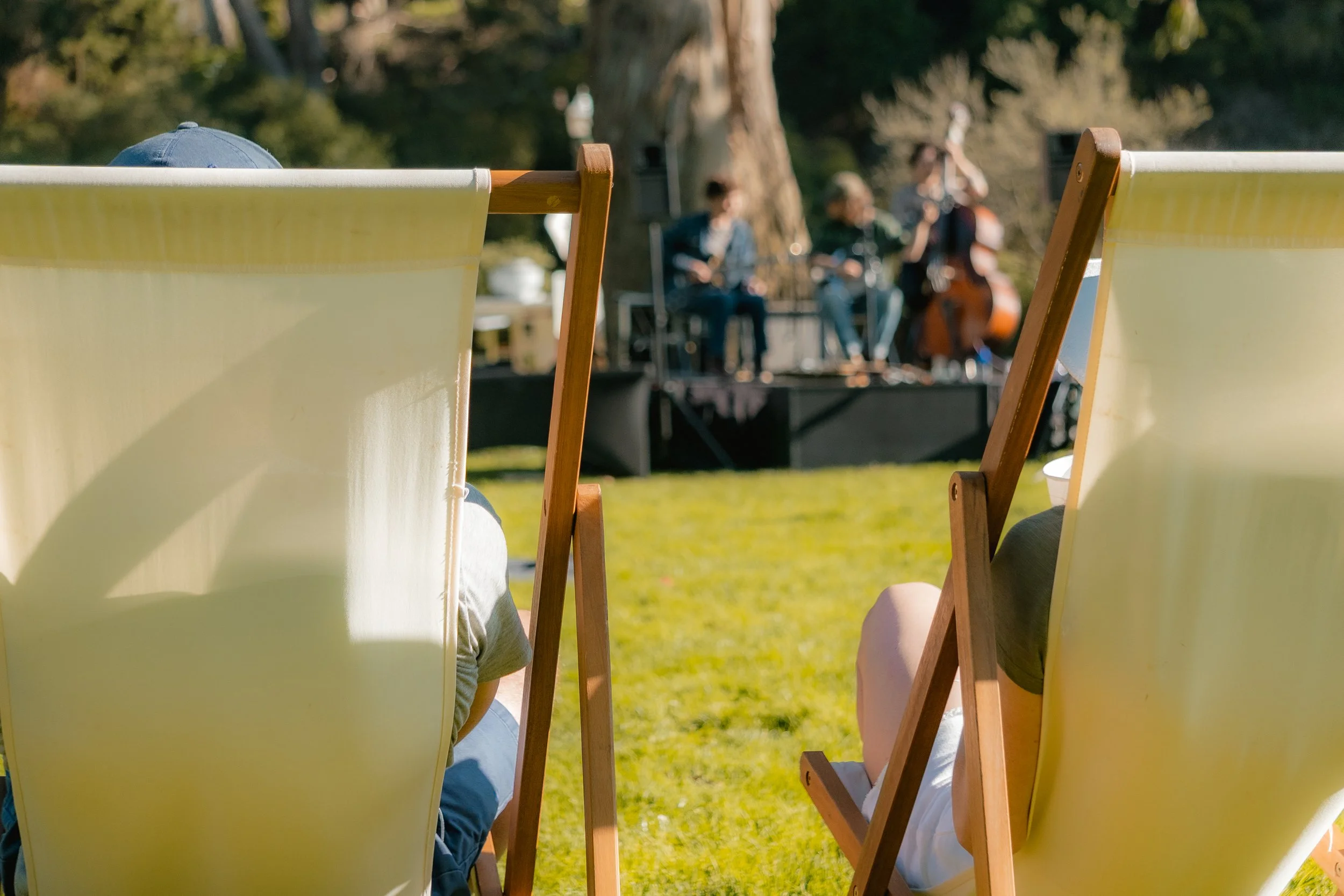People sitting on yellow lounge chairs and watching a band perform on a stage outdoors in a park.