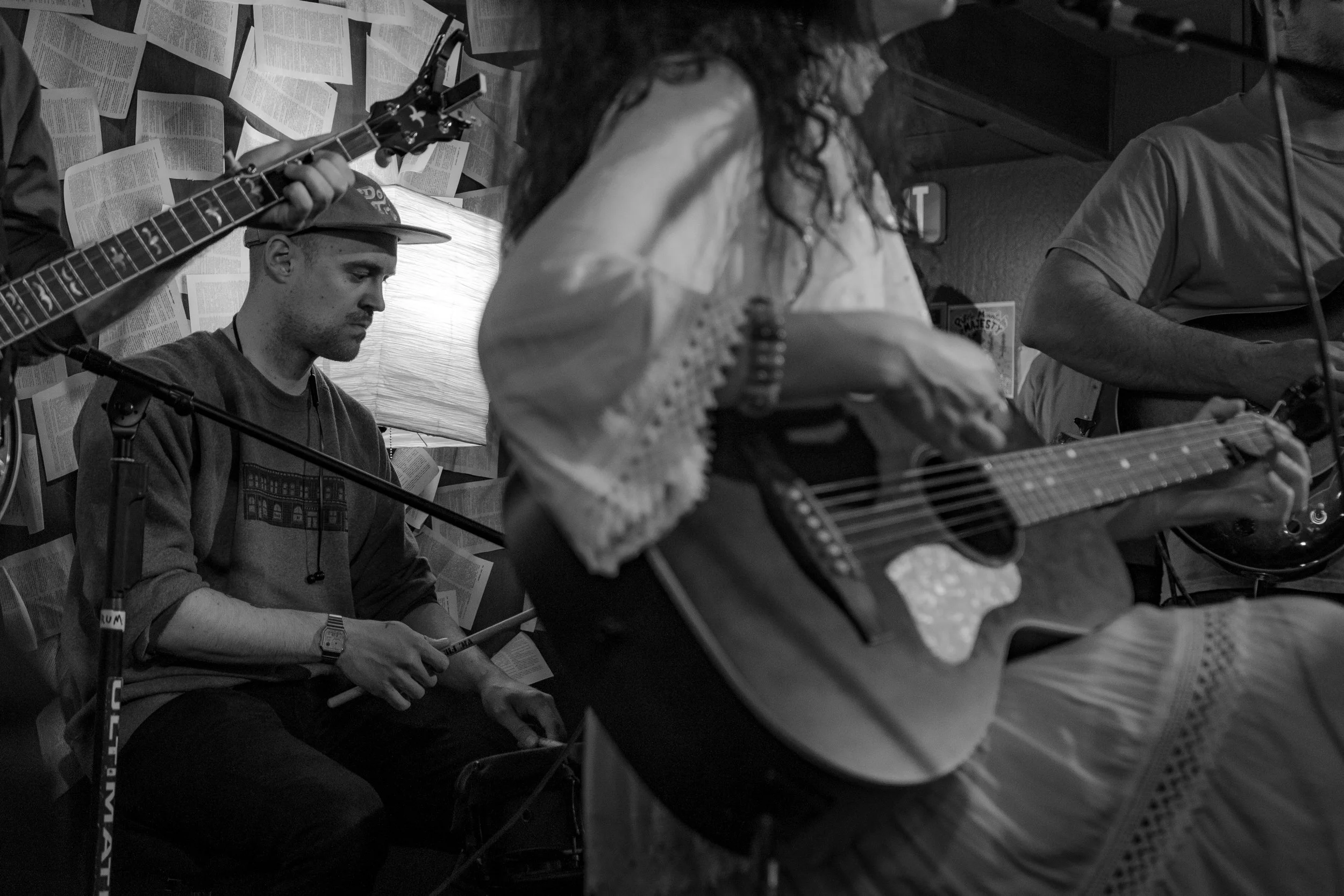 A group of musicians playing instruments in a cozy indoor setting, with sheet music or pages on the wall behind them.