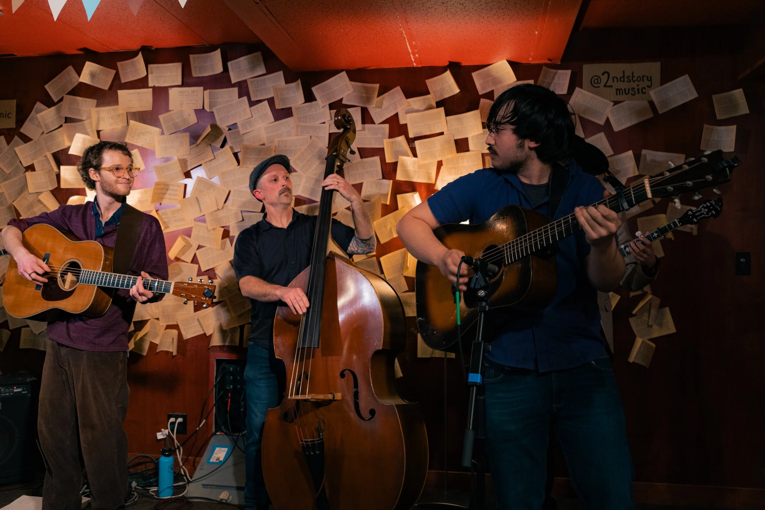 Four musicians playing guitars and a double bass perform on stage against a backdrop decorated with floating pages.