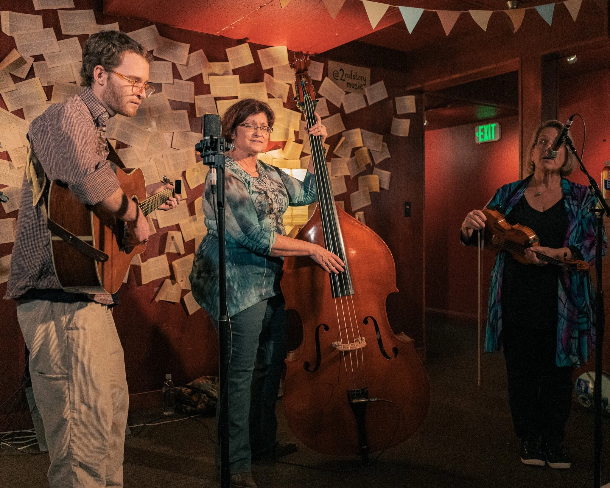 Three musicians performing indoors, two women and one man. The woman on the right sings into a microphone and holds a violin, the woman in the middle plays a double bass, and the man on the left plays an acoustic guitar. Background features pages of 