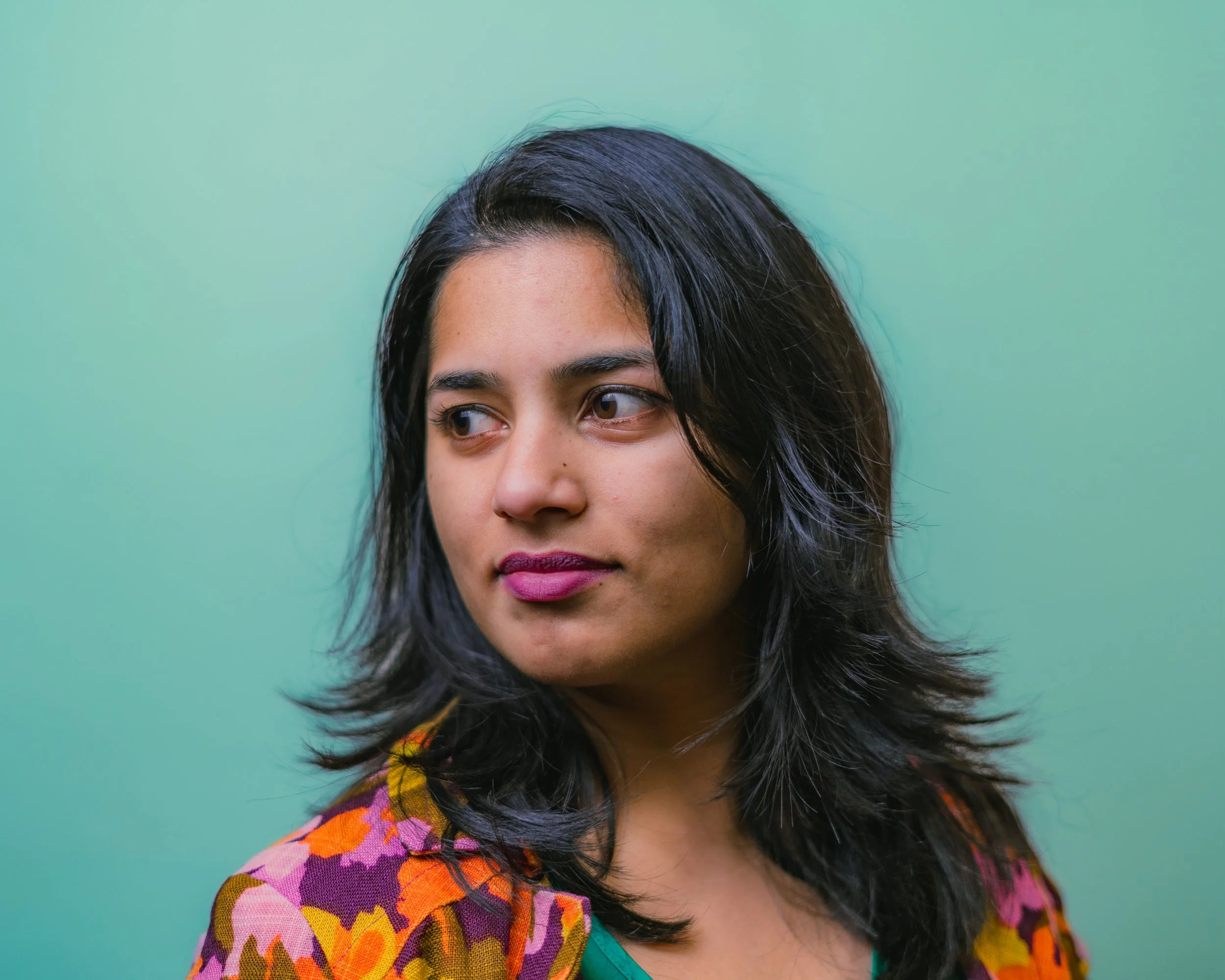 A woman with dark hair wearing a colorful floral-patterned shirt stands against a gradient green background.
