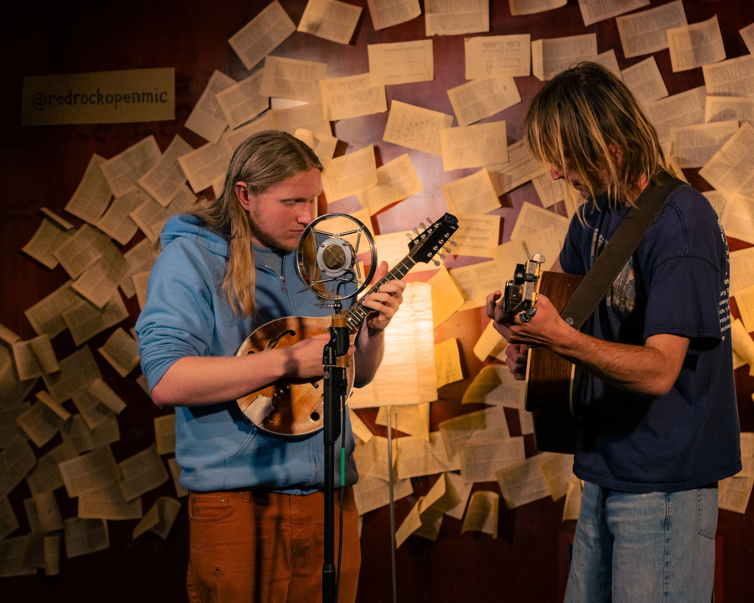 Two young men playing acoustic guitars in a music studio with a vintage microphone and a backdrop of scattered books on the wall.