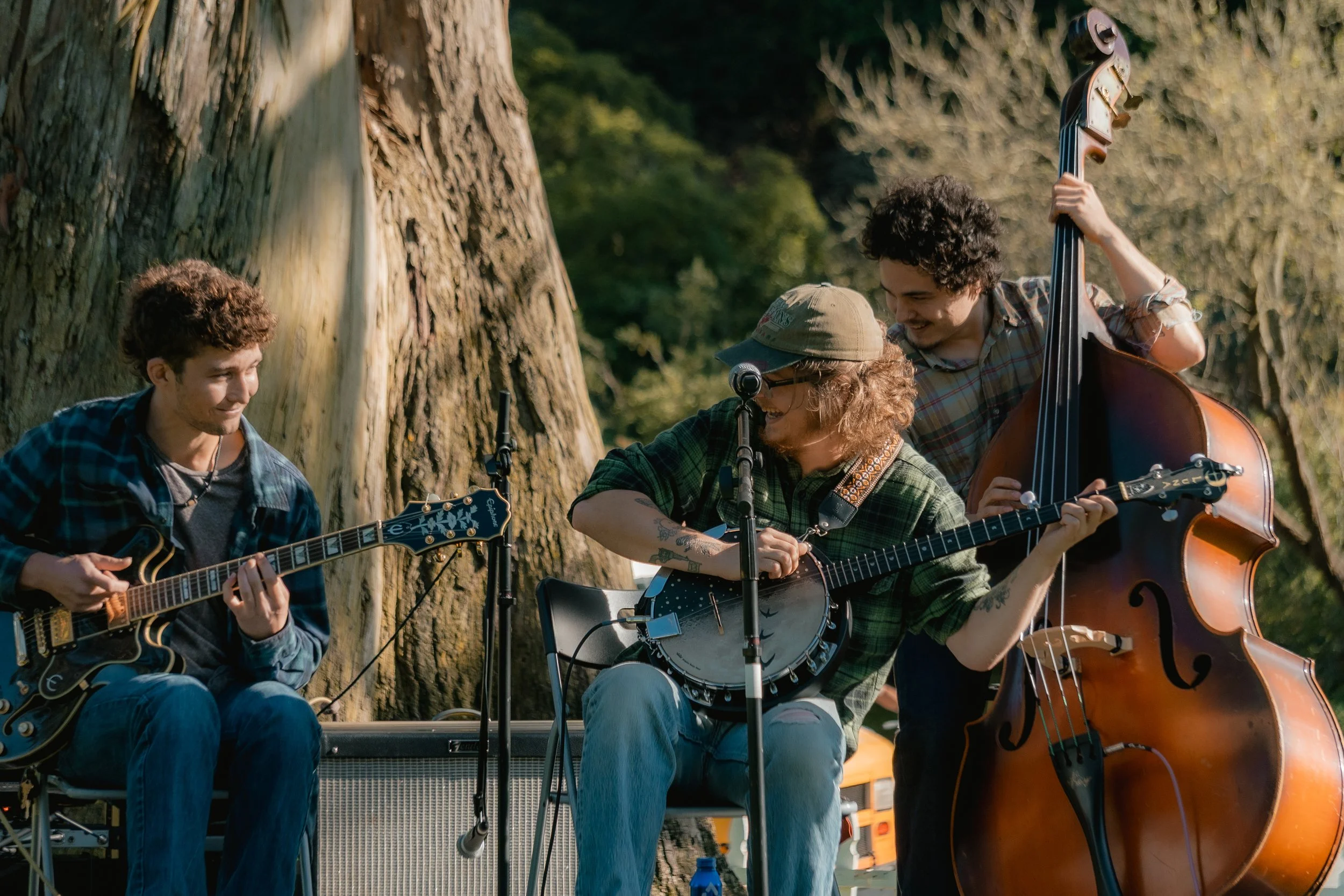 Three young men playing stringed instruments outdoors near a large tree, smiling and enjoying each other's company.