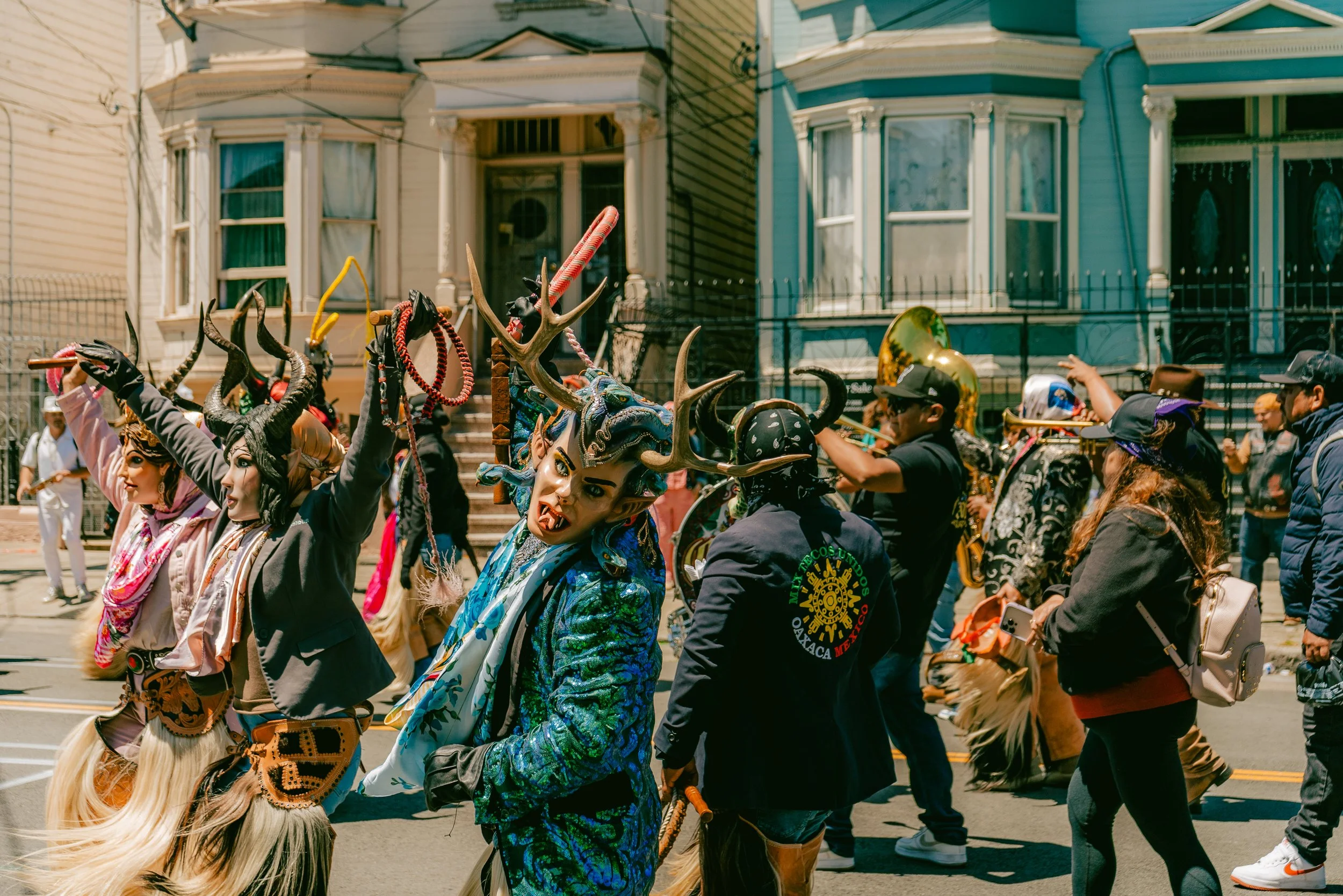 People participating in a parade wearing traditional costumes with horned masks, some playing musical instruments, on a city street with Victorian-style houses in the background.
