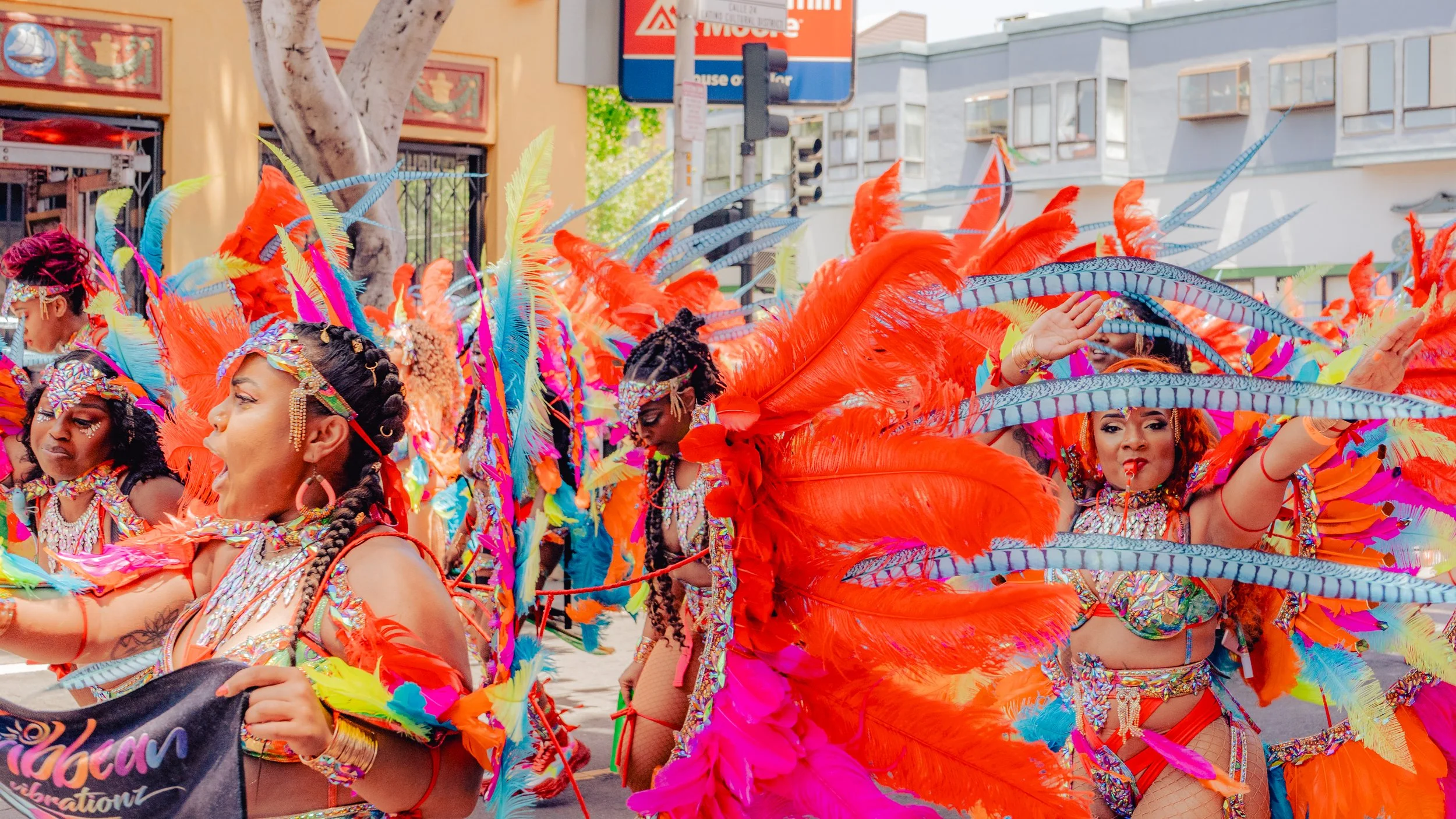 Women in colorful Carnival costumes with feathers and jewelry, participating in a parade on a city street.