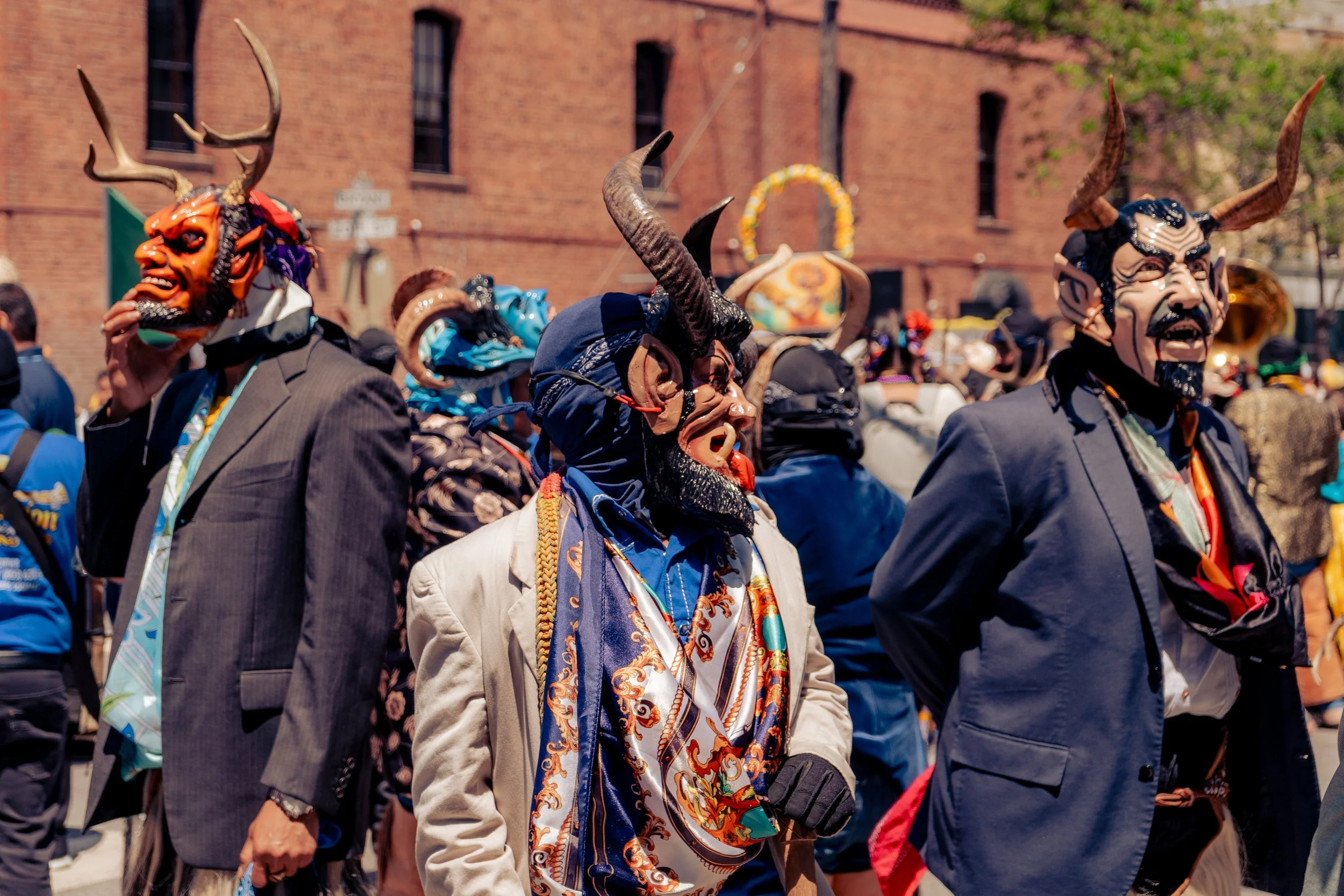 People wearing colorful masks and costumes at a street parade or festival with a brick building in the background.