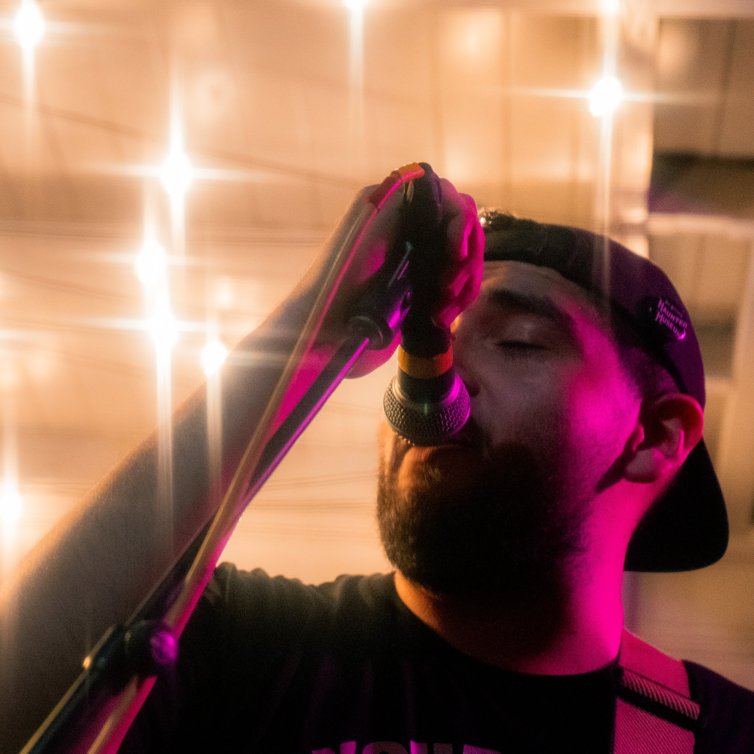 A man singing into a microphone with pink and purple lighting, wearing a black baseball cap backwards and a black shirt, with a beard and a focused expression.