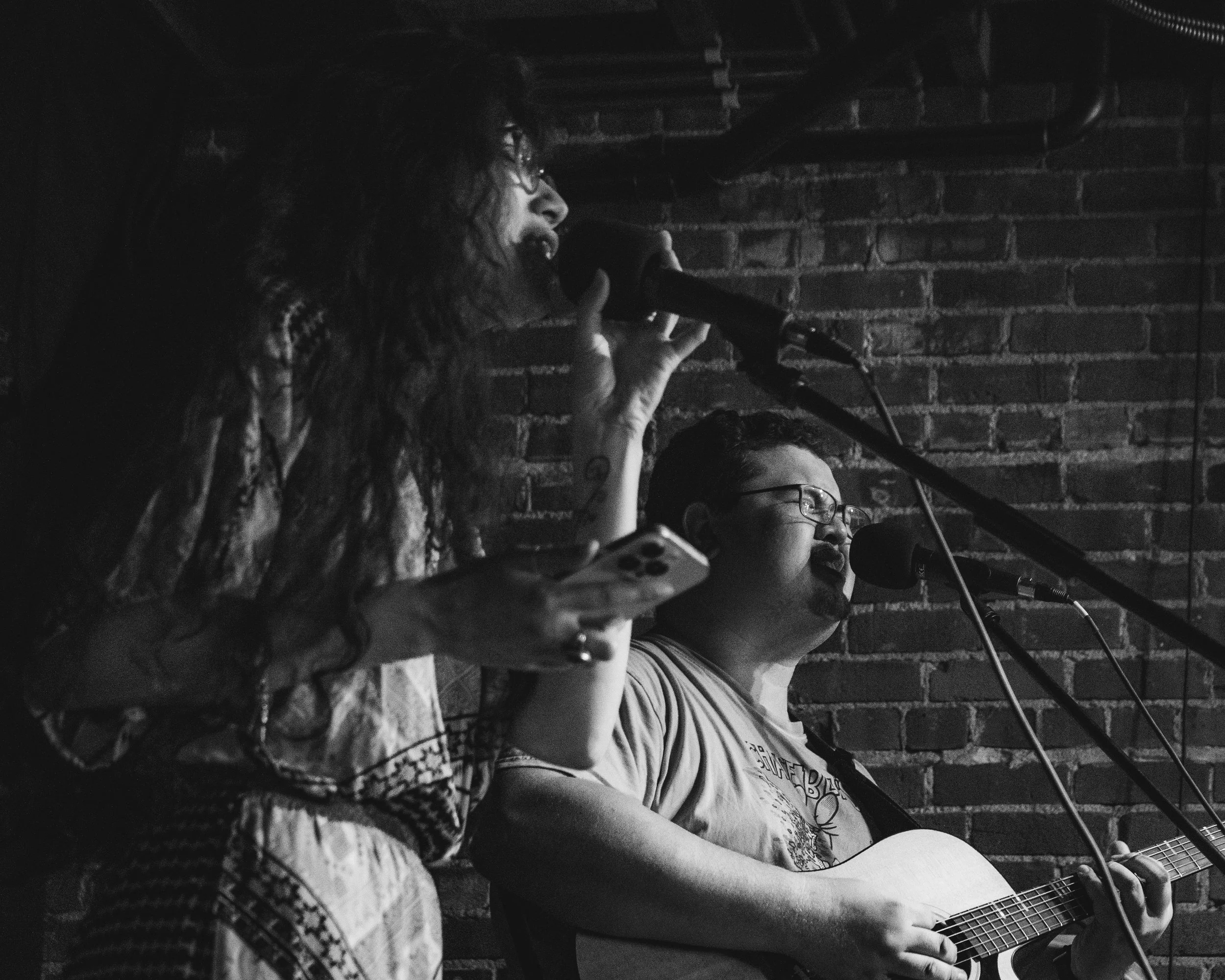 A woman with long curly hair, glasses, and patterned sleeves sings into a microphone while holding a smartphone in one hand. A man with glasses playing an acoustic guitar sings into a microphone. They are performing against a brick wall in a dimly li