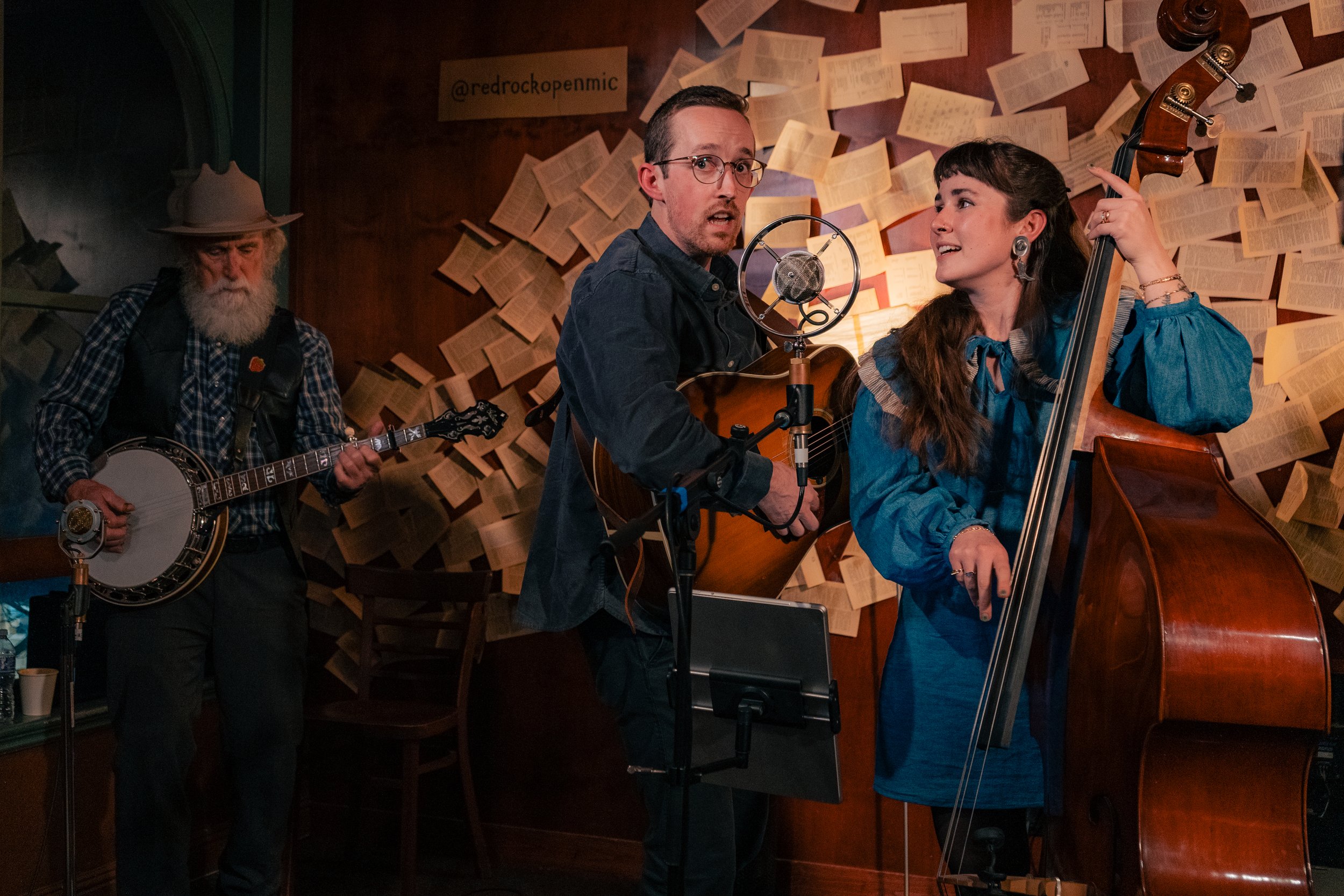 Three musicians performing in a cozy, book-lined venue. One man plays a guitar, another man, with glasses, sings into a mic, and a woman plays a double bass, all focused on their music.
