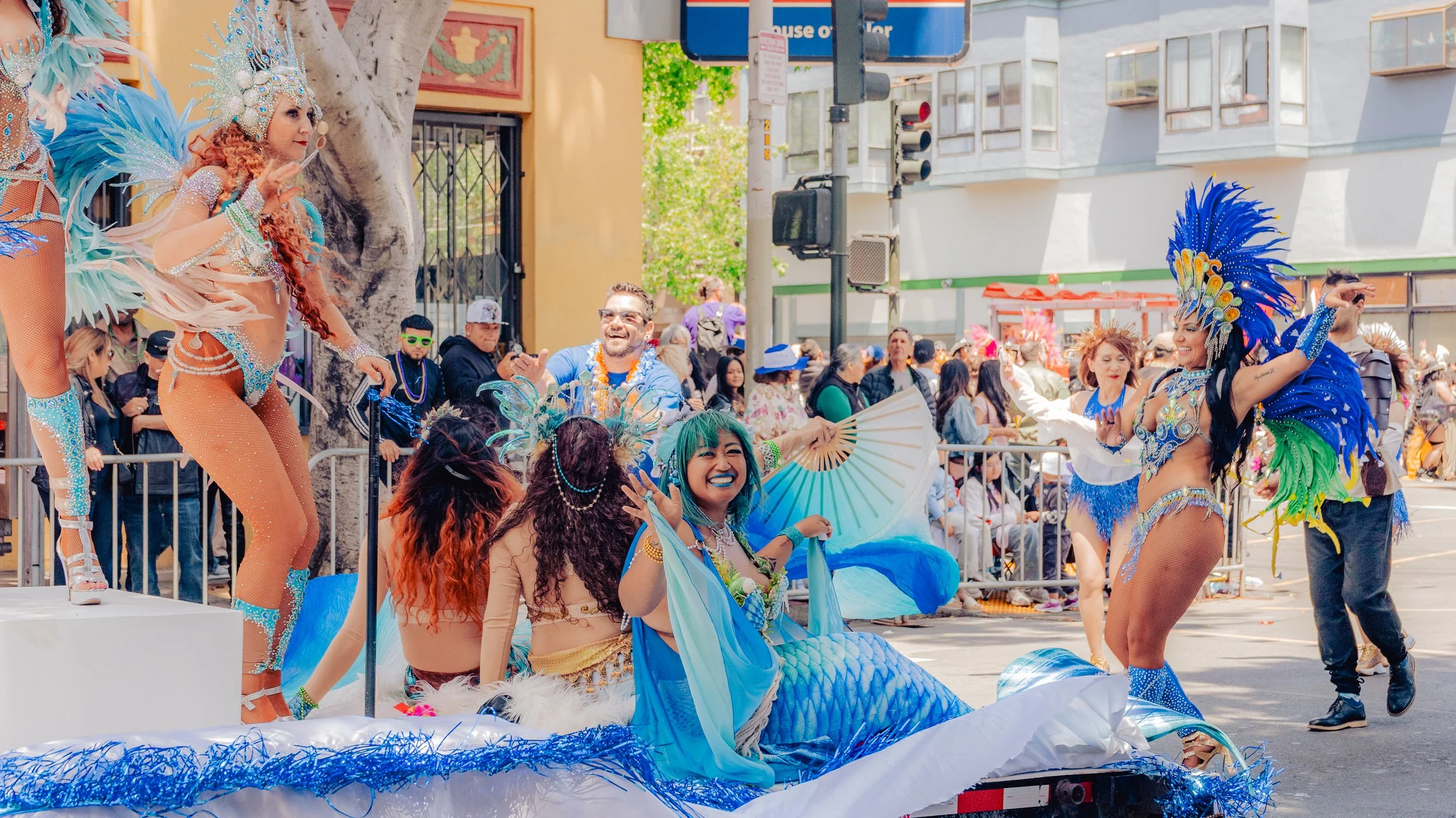 Dancers dressed as mermaids and fantasy characters parade and dance during a street festival, with spectators on the sidelines and buildings in the background.