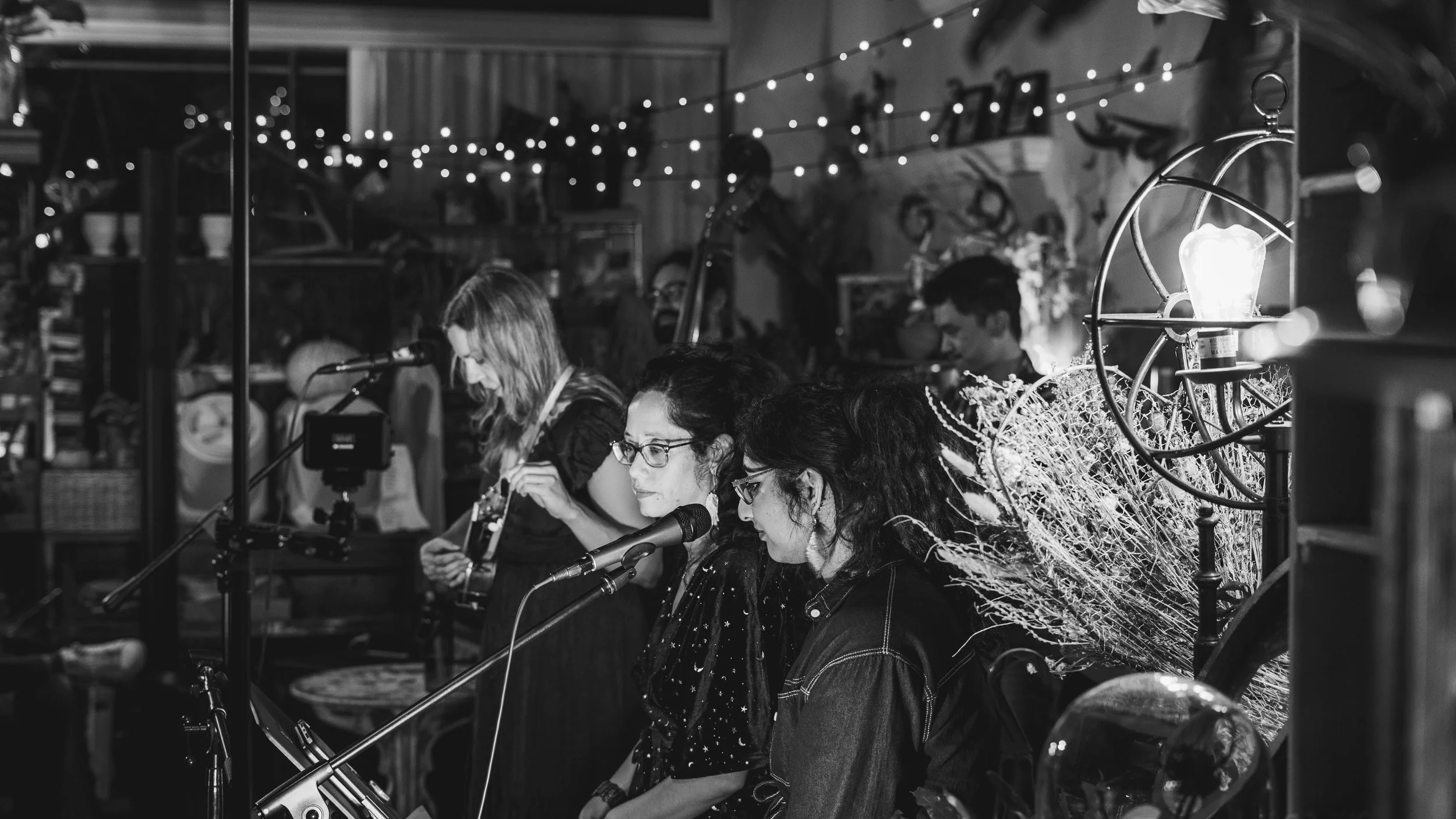 Black and white photo of a live music performance in a cozy, decorated space. Four women are singing or playing instruments, with some wearing glasses. One woman is playing a guitar, and another is singing into a microphone. The background shows shel