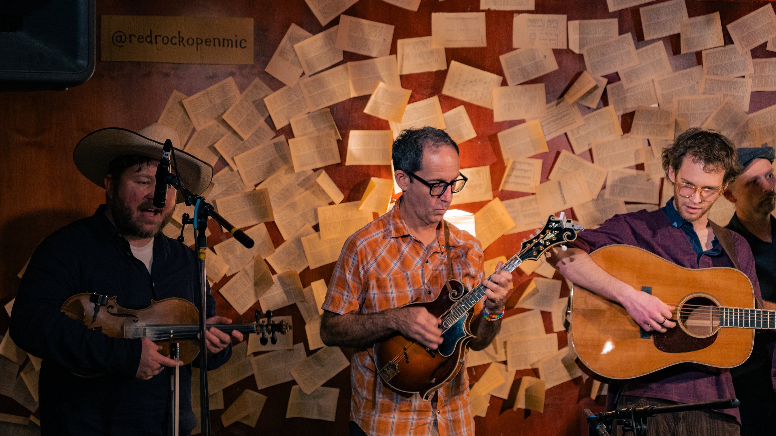 Four musicians performing with string instruments in front of a backdrop decorated with open books