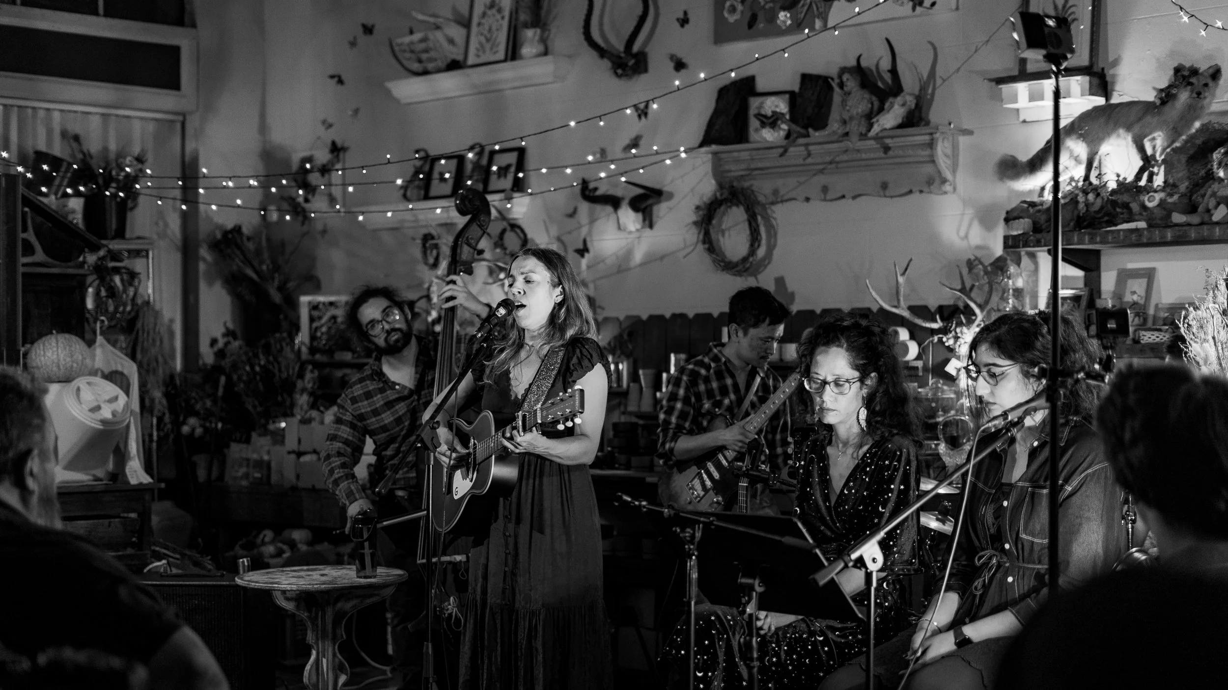A black and white photo of a live music performance in an intimate indoor setting. A woman in a dress is singing and playing an acoustic guitar into a microphone, surrounded by other musicians, including violinists. The background has decorative anim