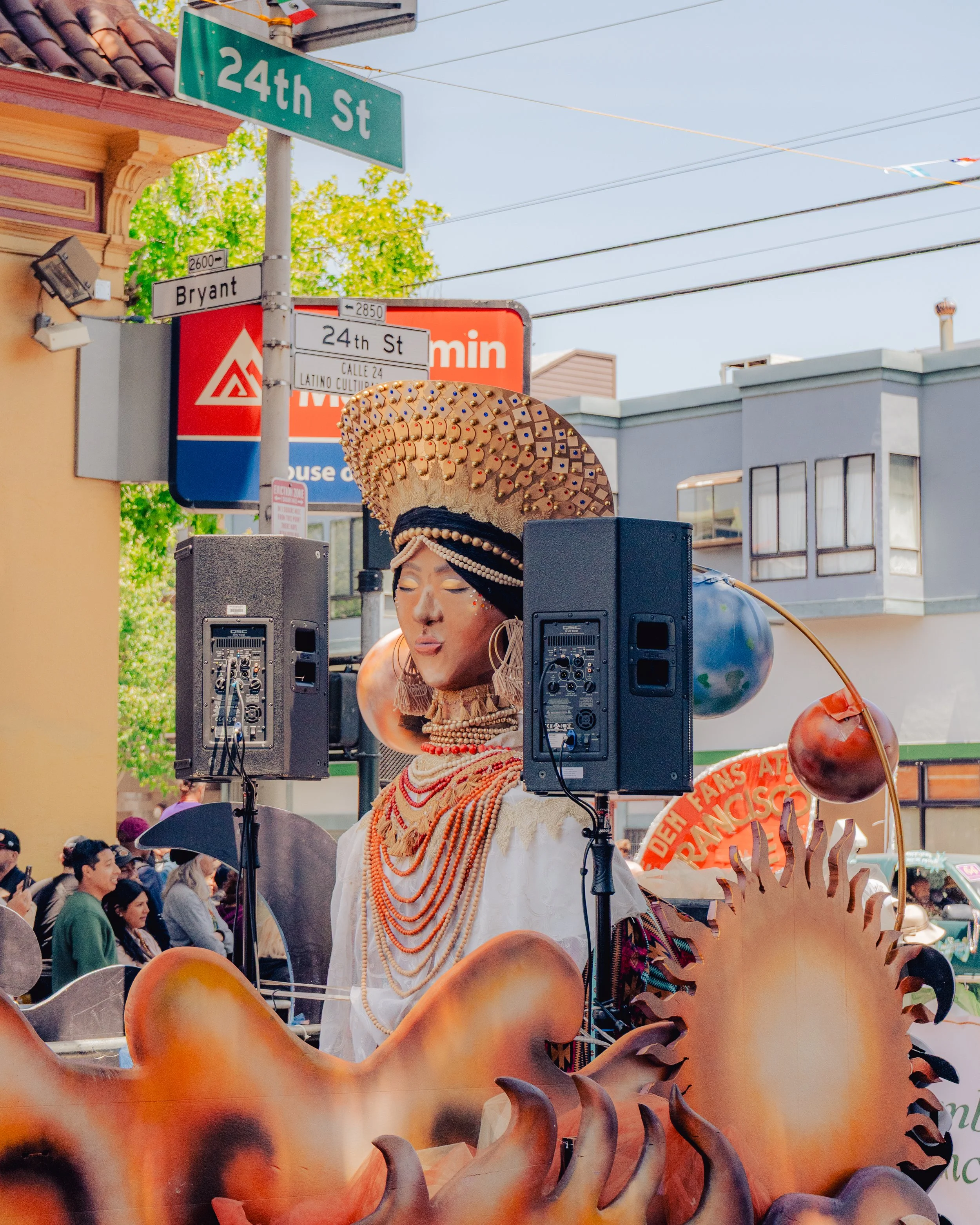 A person dressed in traditional Indigenous attire with a large decorated headdress, jewelry, and face paint, standing among speakers and decorations at a street parade on 24th Street.
