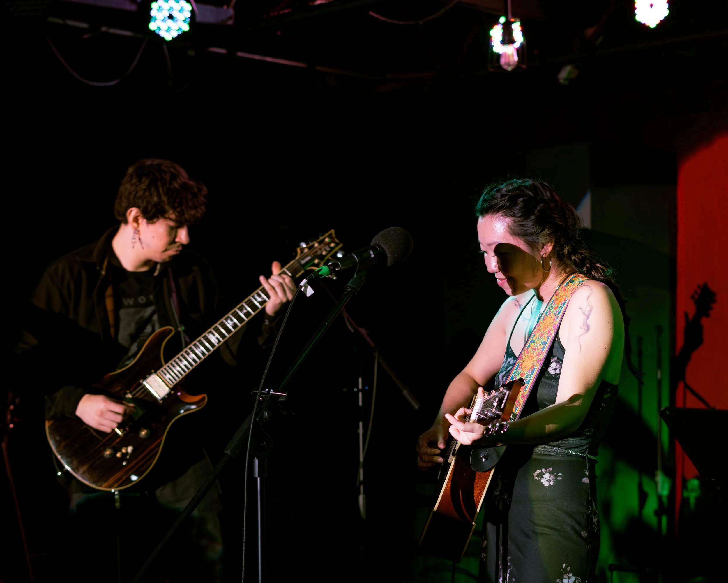 Two musicians playing guitars on stage, a woman on the right and a man on the left, with a microphone between them, in a dimly lit venue with colorful lighting.