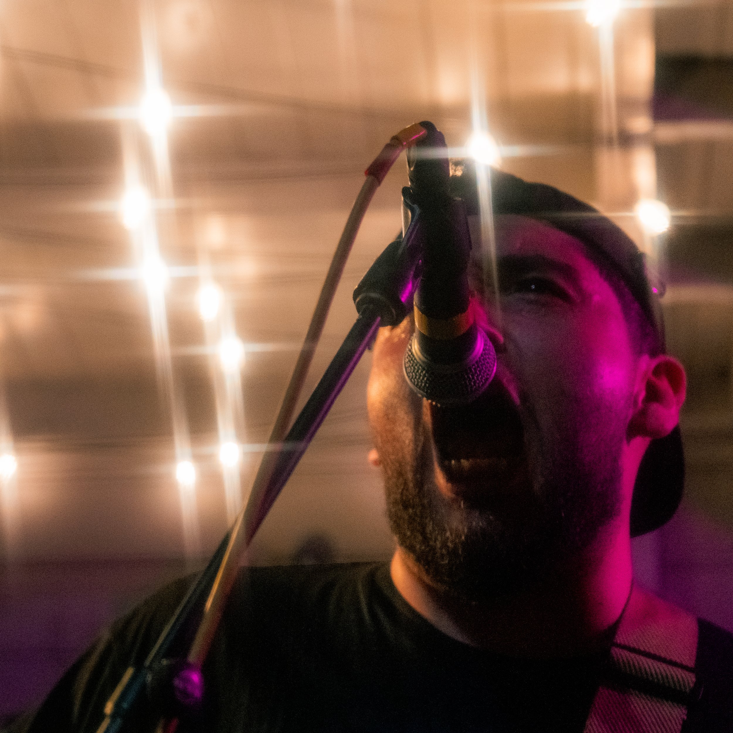 A man singing into a microphone with a microphone stand, illuminated by purple and yellow stage lighting, with a background of bright star-like lights.