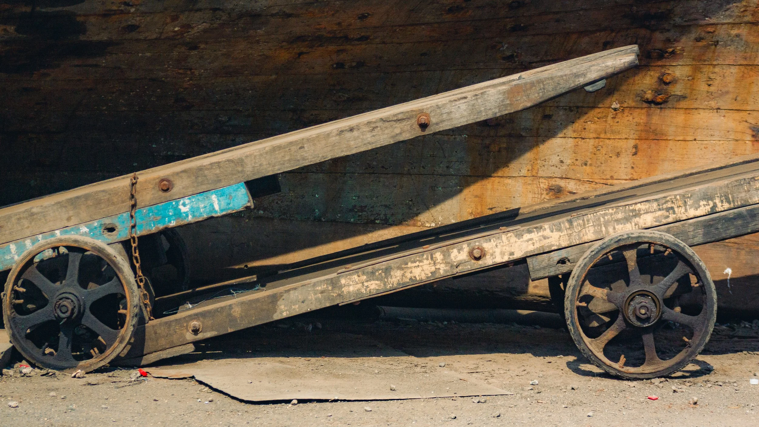Old, weathered wooden dolly with metal wheels, leaning against a wooden wall.