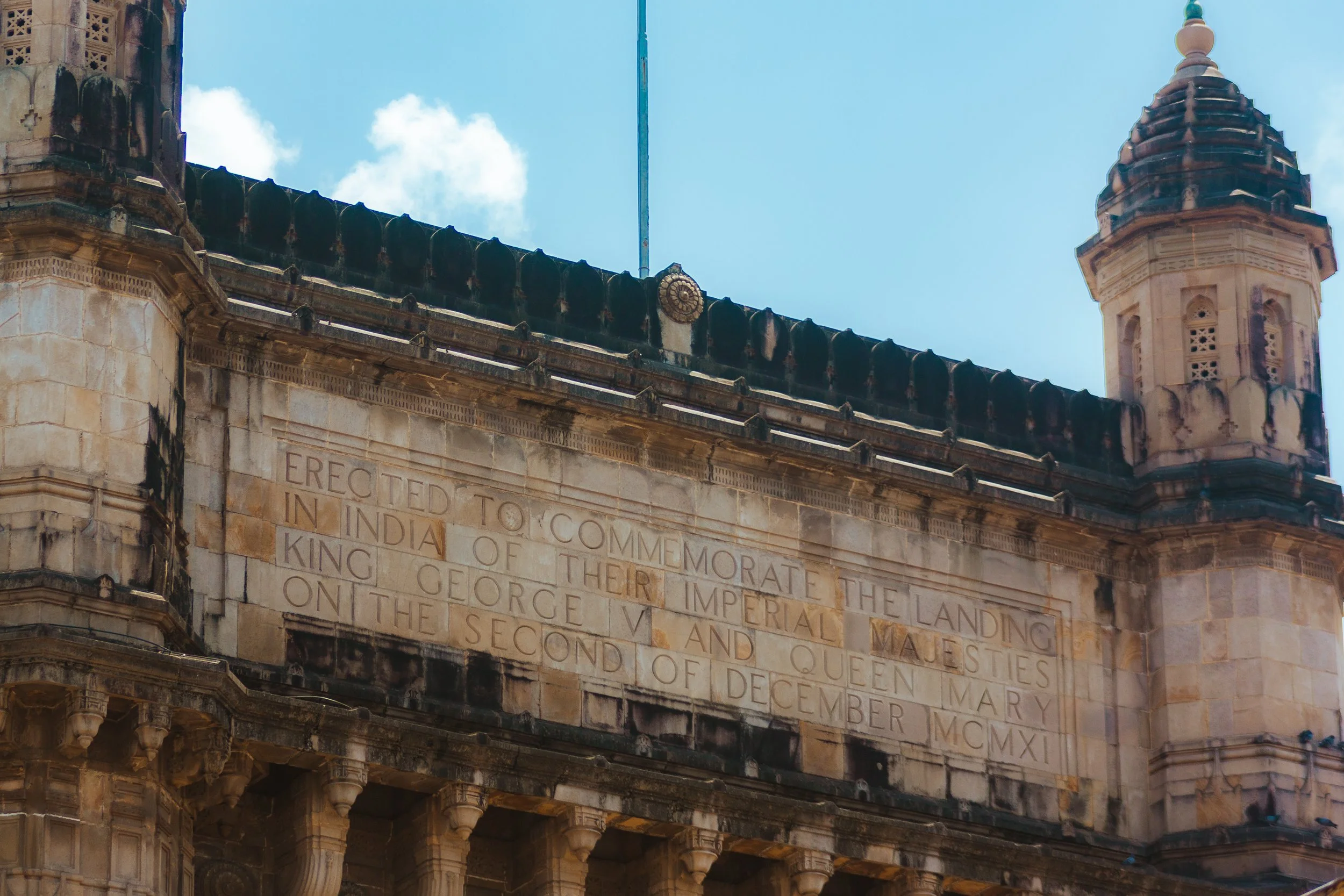 Close-up of an old stone building with an engraved plaque, a tower, and a blue sky with some clouds. The plaque commemorates the landing of King George V and Queen Mary in India.