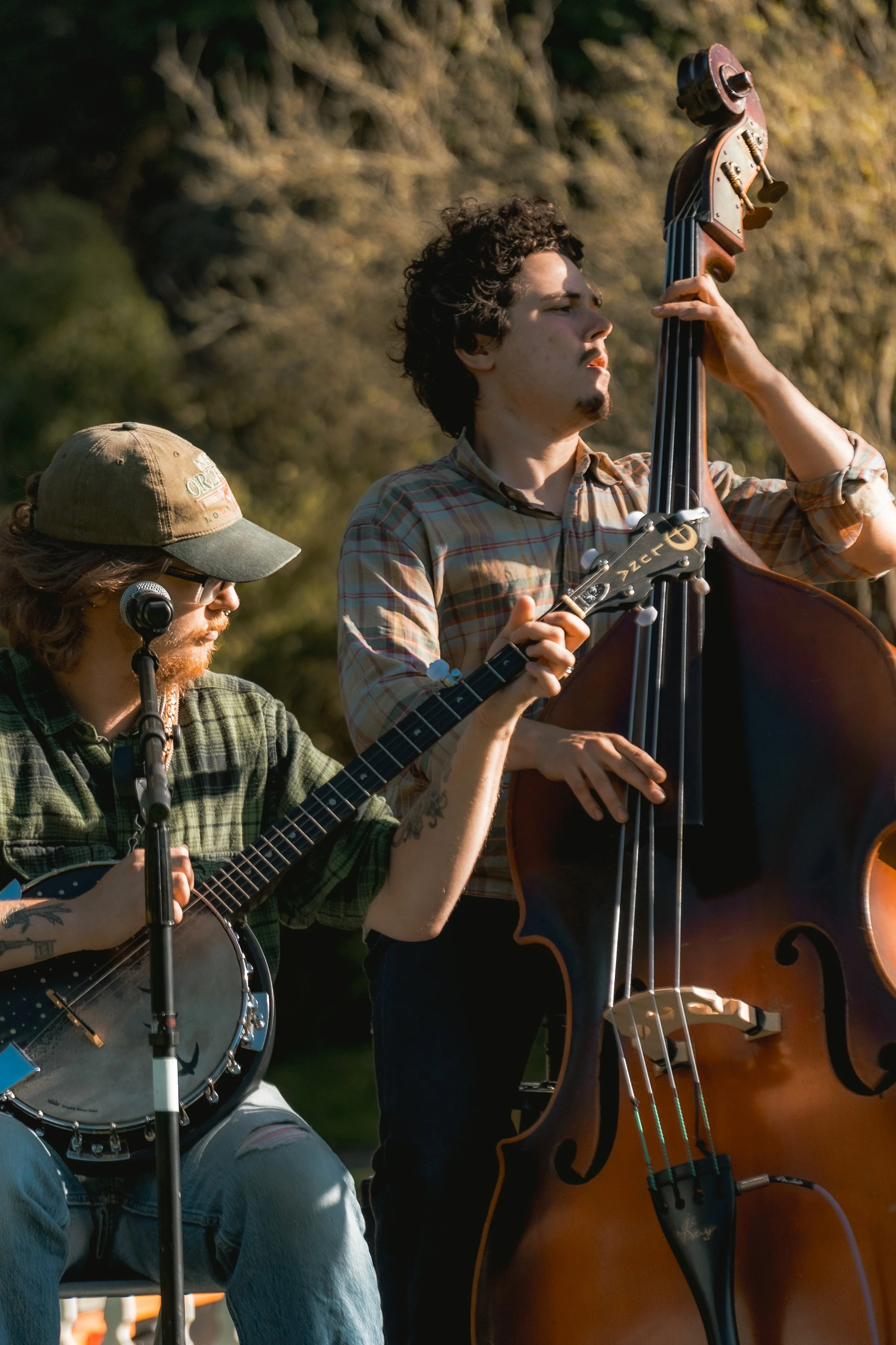 Two musicians playing string instruments outdoors during daylight. The person on the left is playing a banjo, wearing glasses, a cap, and a plaid shirt. The person on the right is playing an upright bass, wearing a plaid shirt, with curly hair, and a