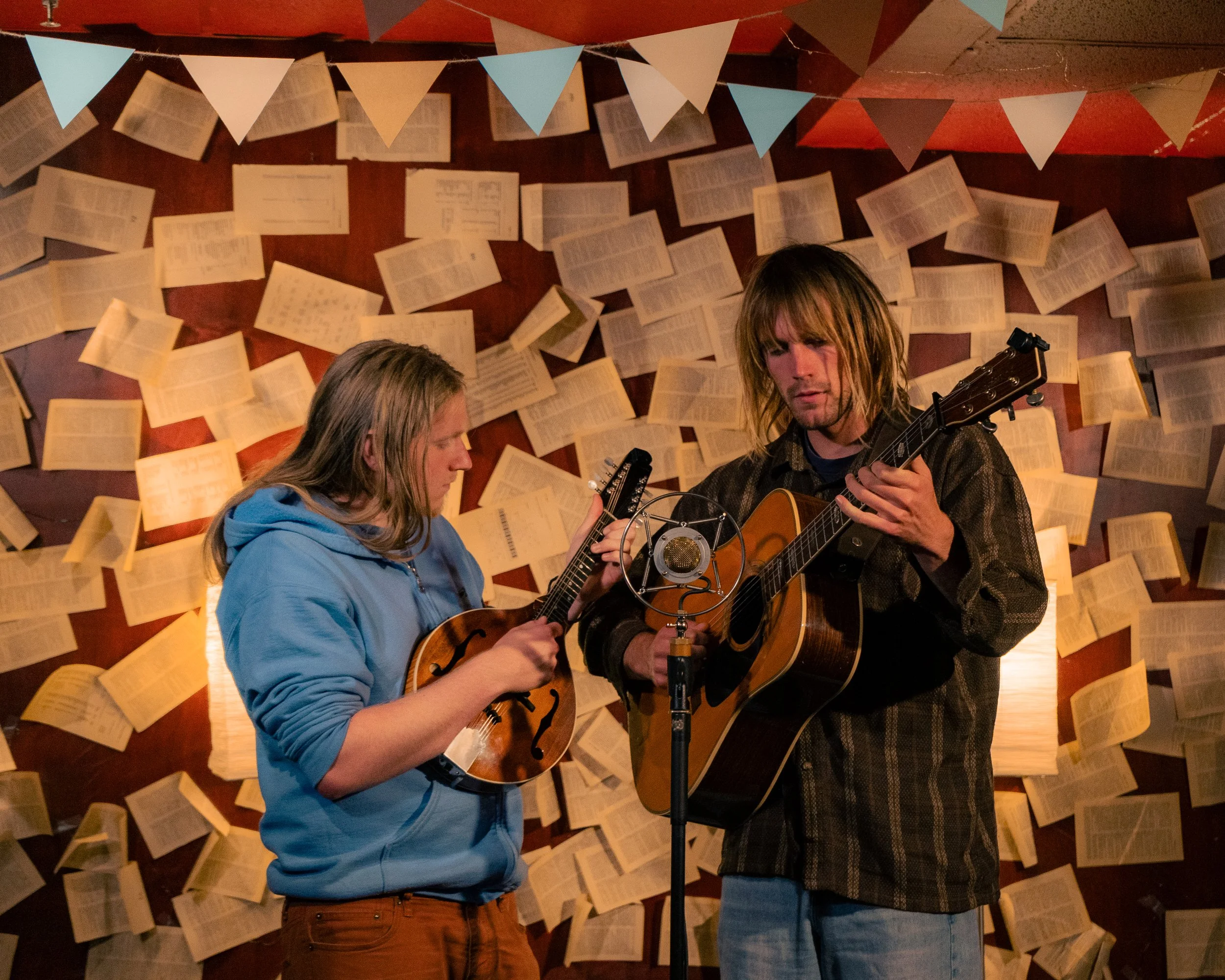 Two musicians playing guitars under a vintage microphone, with a background of open books and paper pages on the wall.