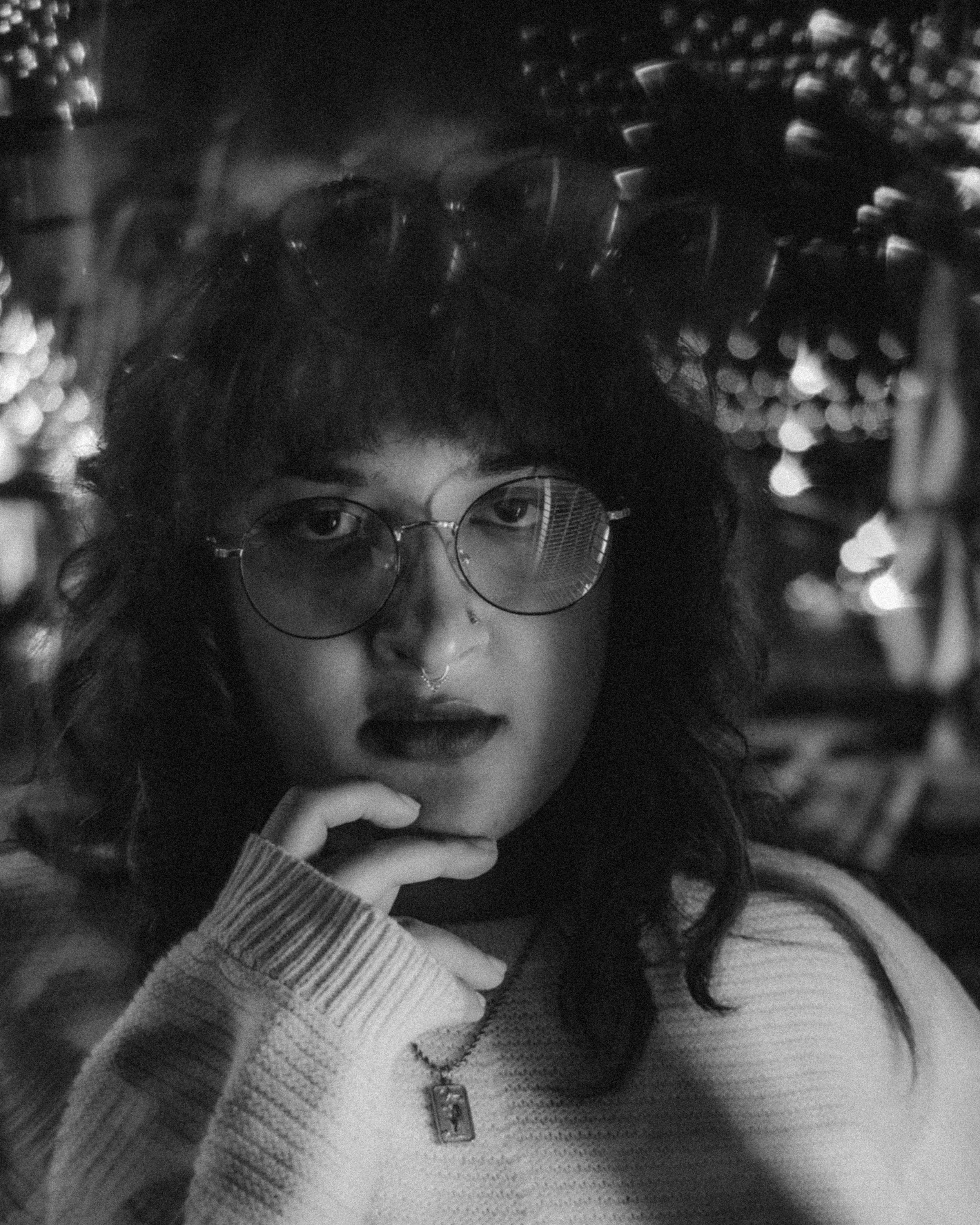 A black and white photo of a woman with curly hair wearing glasses with round lenses, dark lipstick, a septum piercing, and a sweater, resting her chin on her hand with her gaze directed at the camera.
