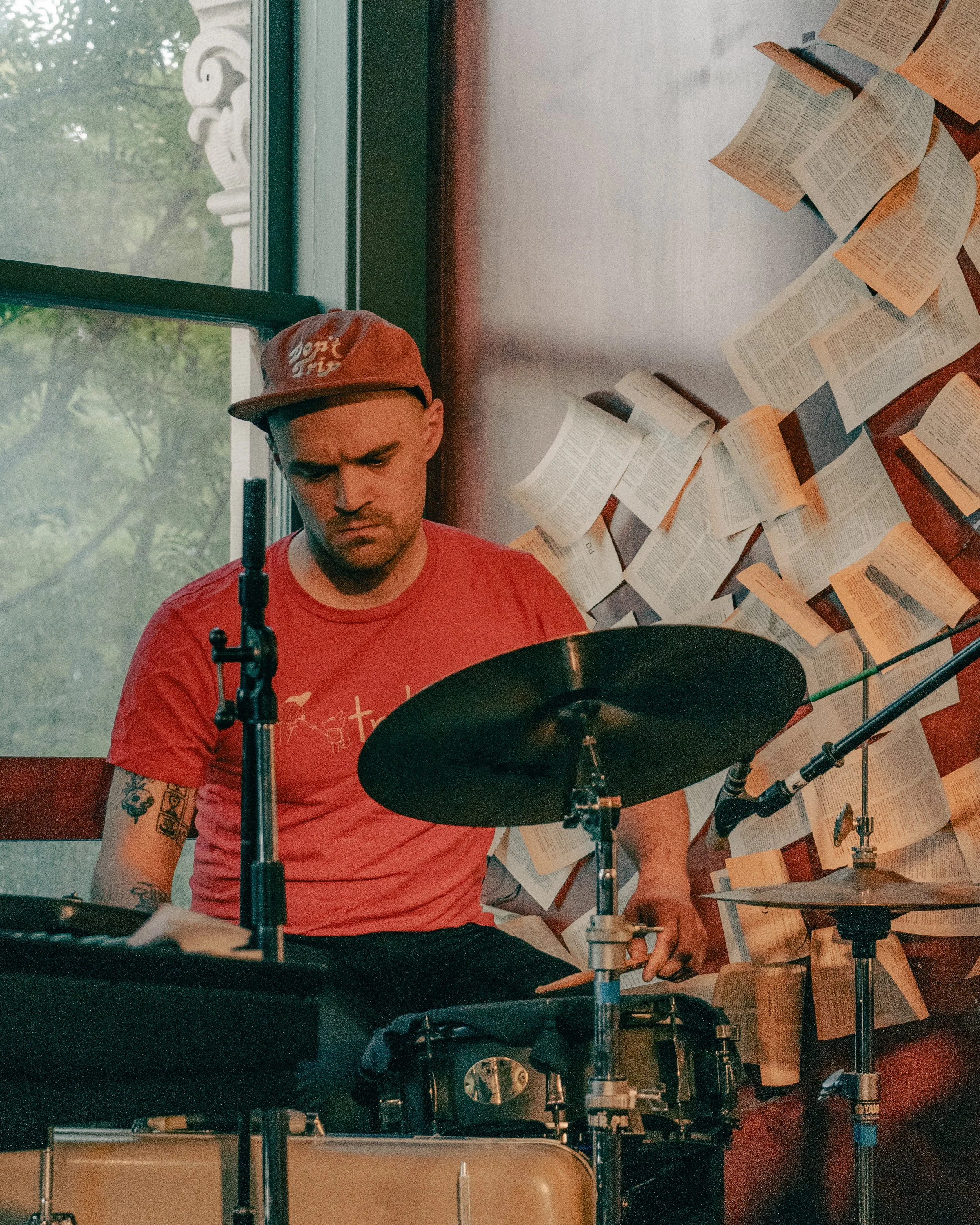 A man wearing a red t-shirt and a baseball cap plays the drums indoors near a window with a wall decorated with open books.