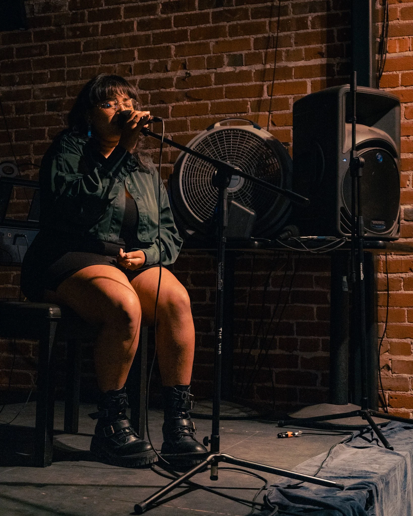 A woman sitting on a chair, singing into a microphone on a stage with brick wall background, wearing glasses, a green jacket, and black combat boots.