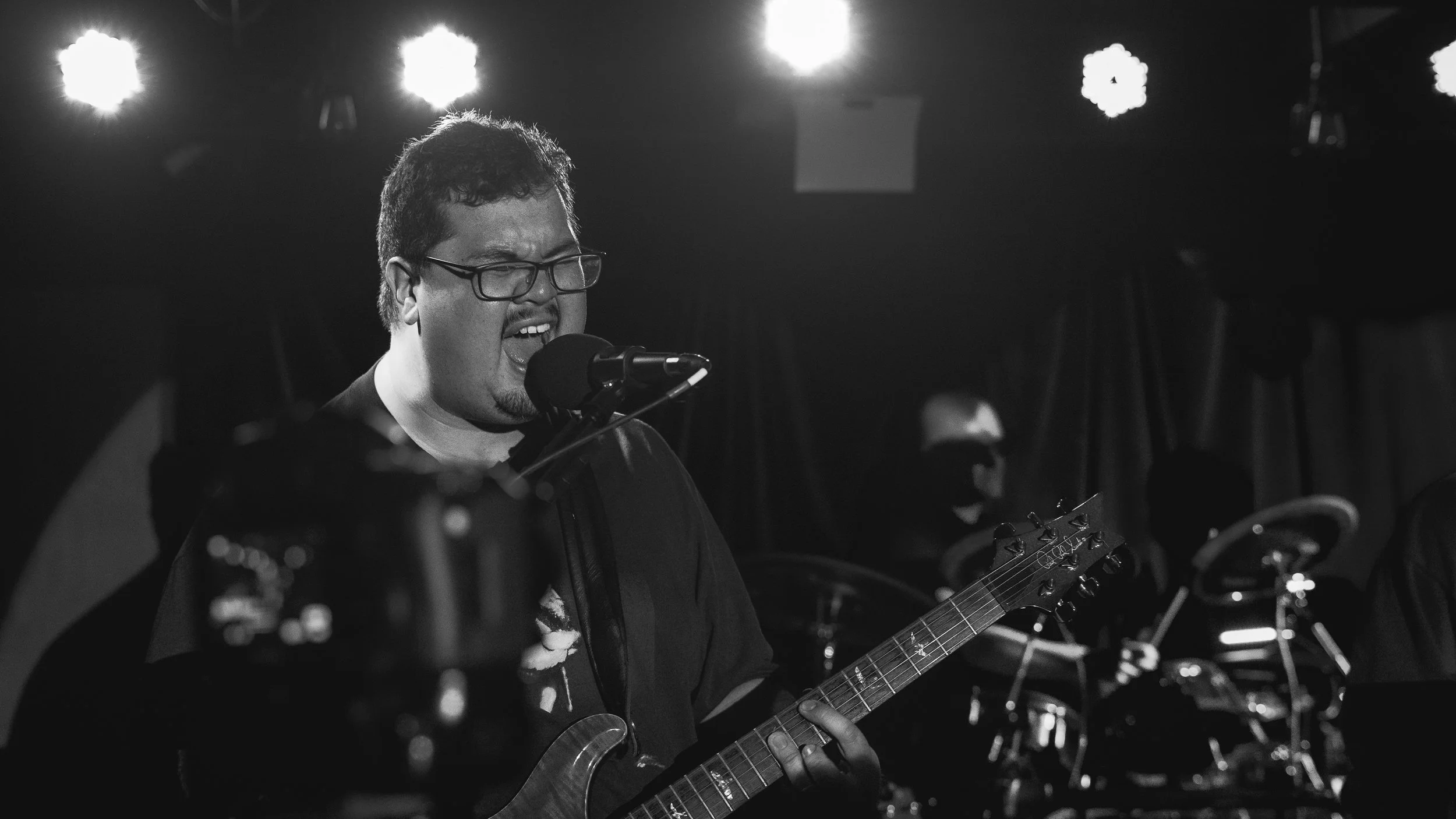 A man wearing glasses singing into a microphone while playing an electric guitar on stage, with a person playing drums in the background, under stage lights.