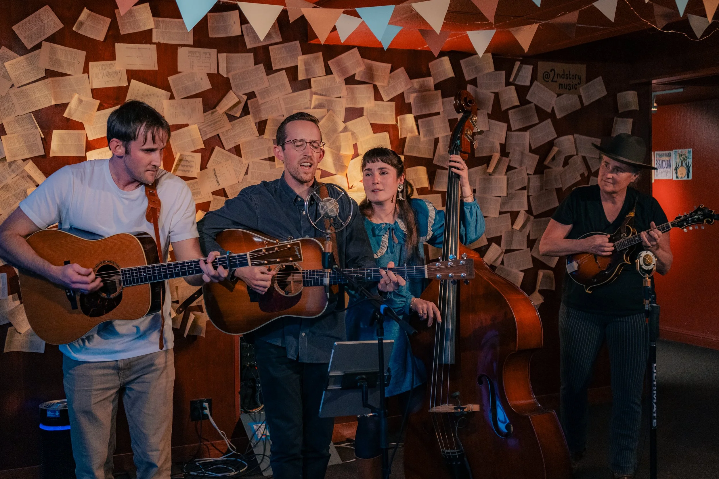 Four musicians performing with guitars and a double bass on a stage decorated with hanging book pages and paper flags.