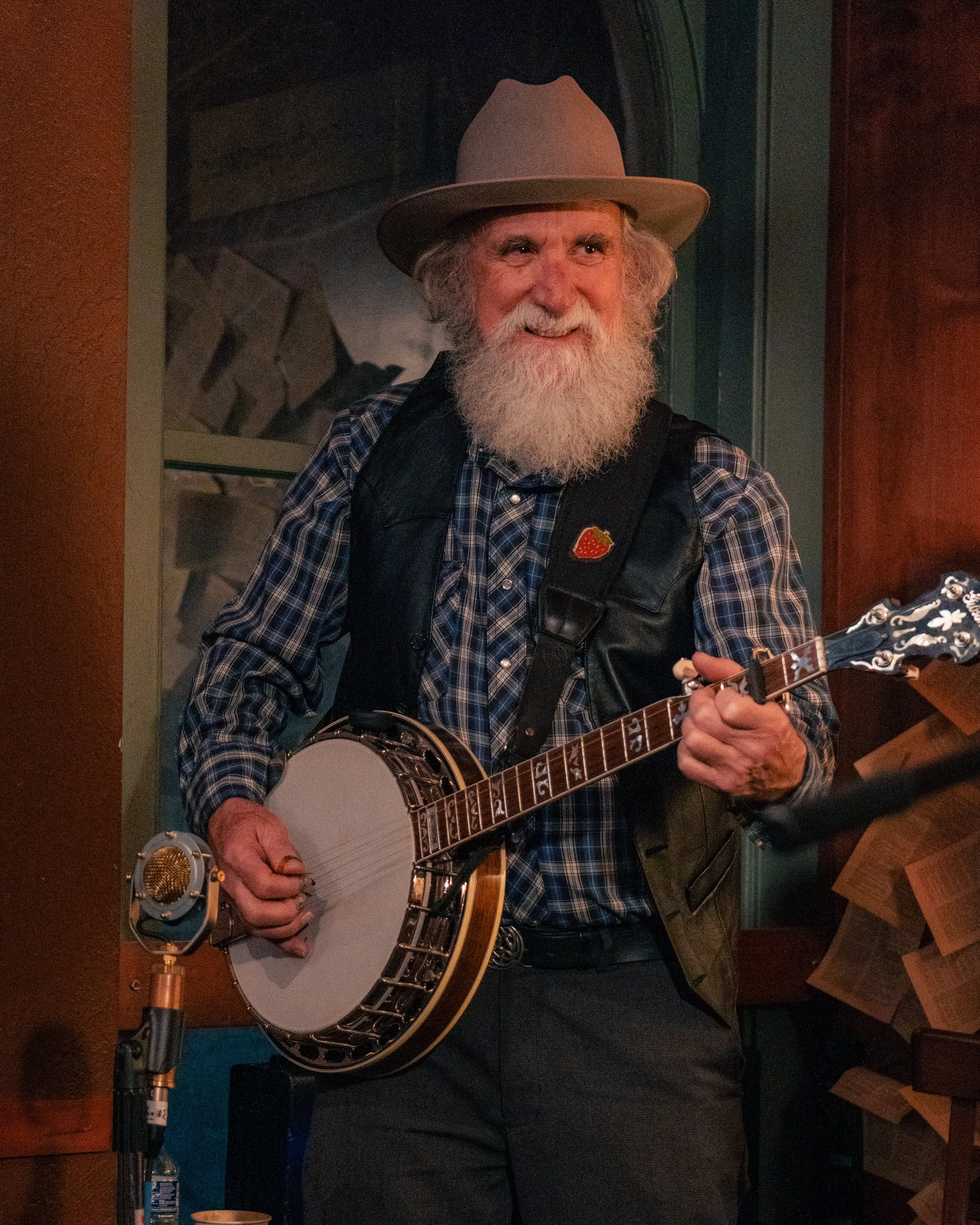 An elderly man with a white beard wearing a beige hat, plaid shirt, and black vest, playing a banjo inside a room.