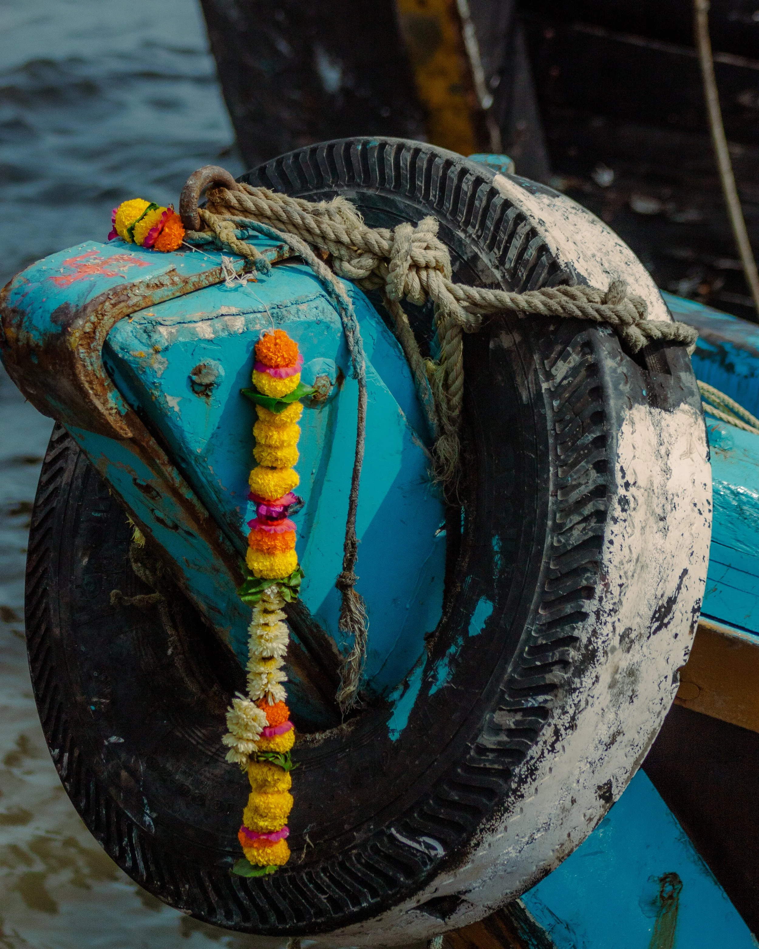 Close-up of a weathered boat wheel attached to a blue boat, decorated with colorful flower garlands and tied with a thick rope, with water in the background.