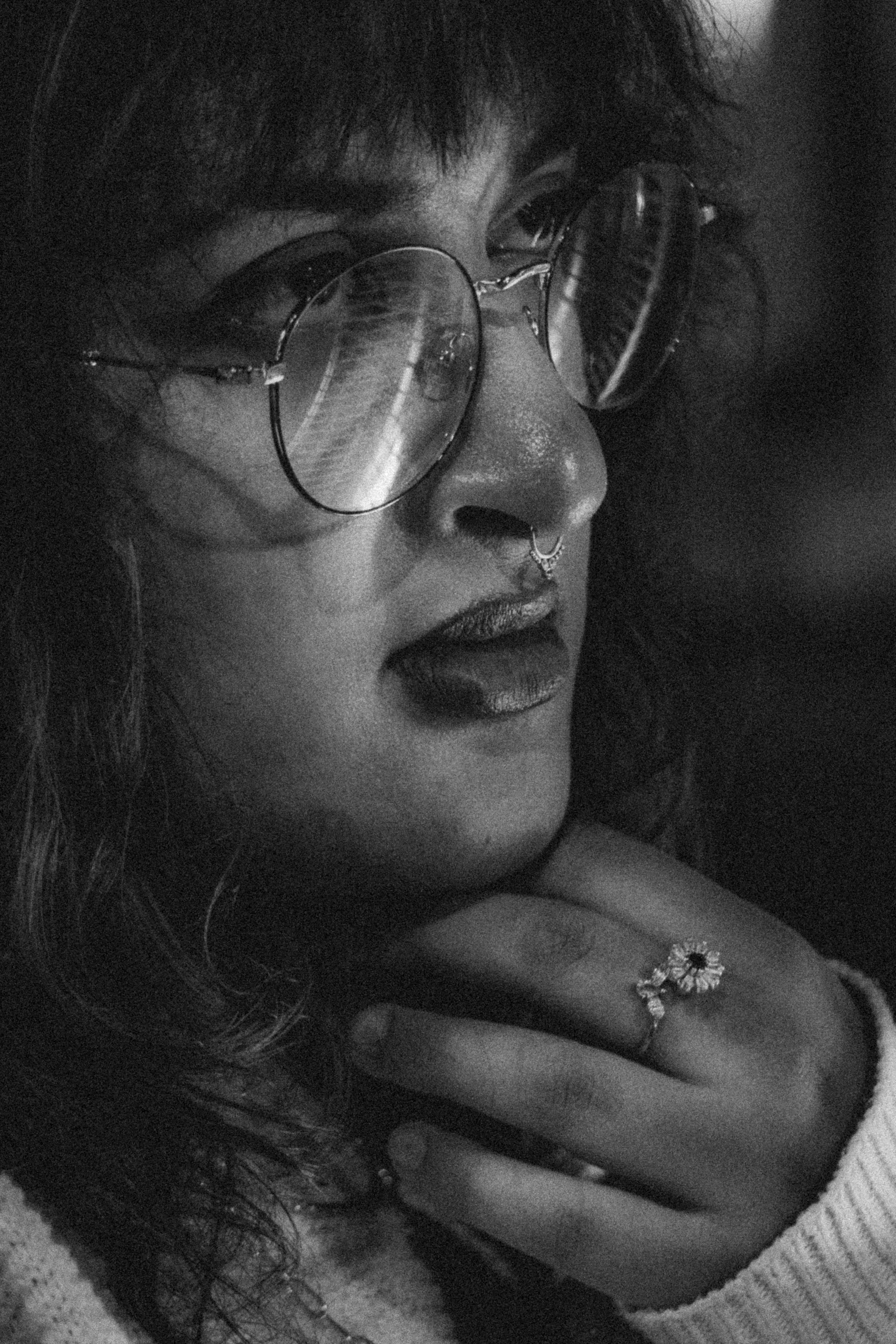 A black-and-white close-up portrait of a woman wearing round glasses, with a septum piercing, lipstick, and a ring on her finger. Her hair is curly and she is resting her chin on her hand.