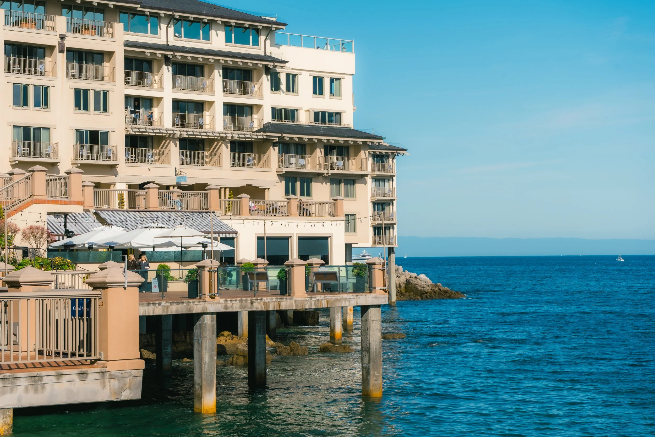 A multi-story beachfront hotel or apartment building with balconies overlooking the ocean, with a patio area with umbrellas and people near the water, and a boat sailing in the distance on a sunny day.