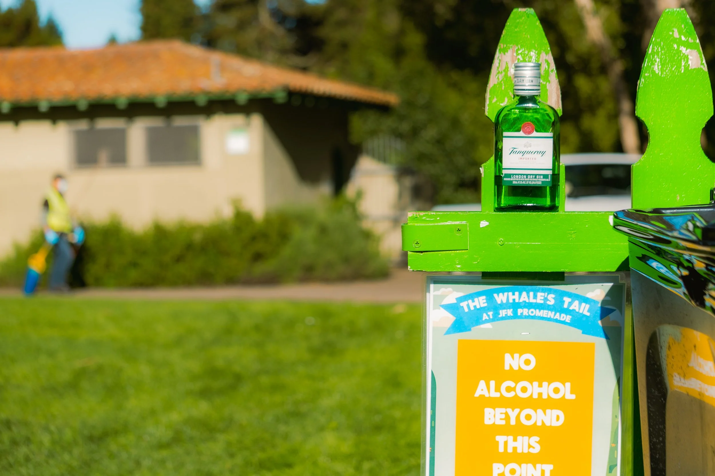 A sign on a green stand that reads, "No alcohol beyond this point," next to a small bottle of Tanqueray London Dry Gin at the JFK Promenade, with a blurred background of a person in a yellow vest and a building.