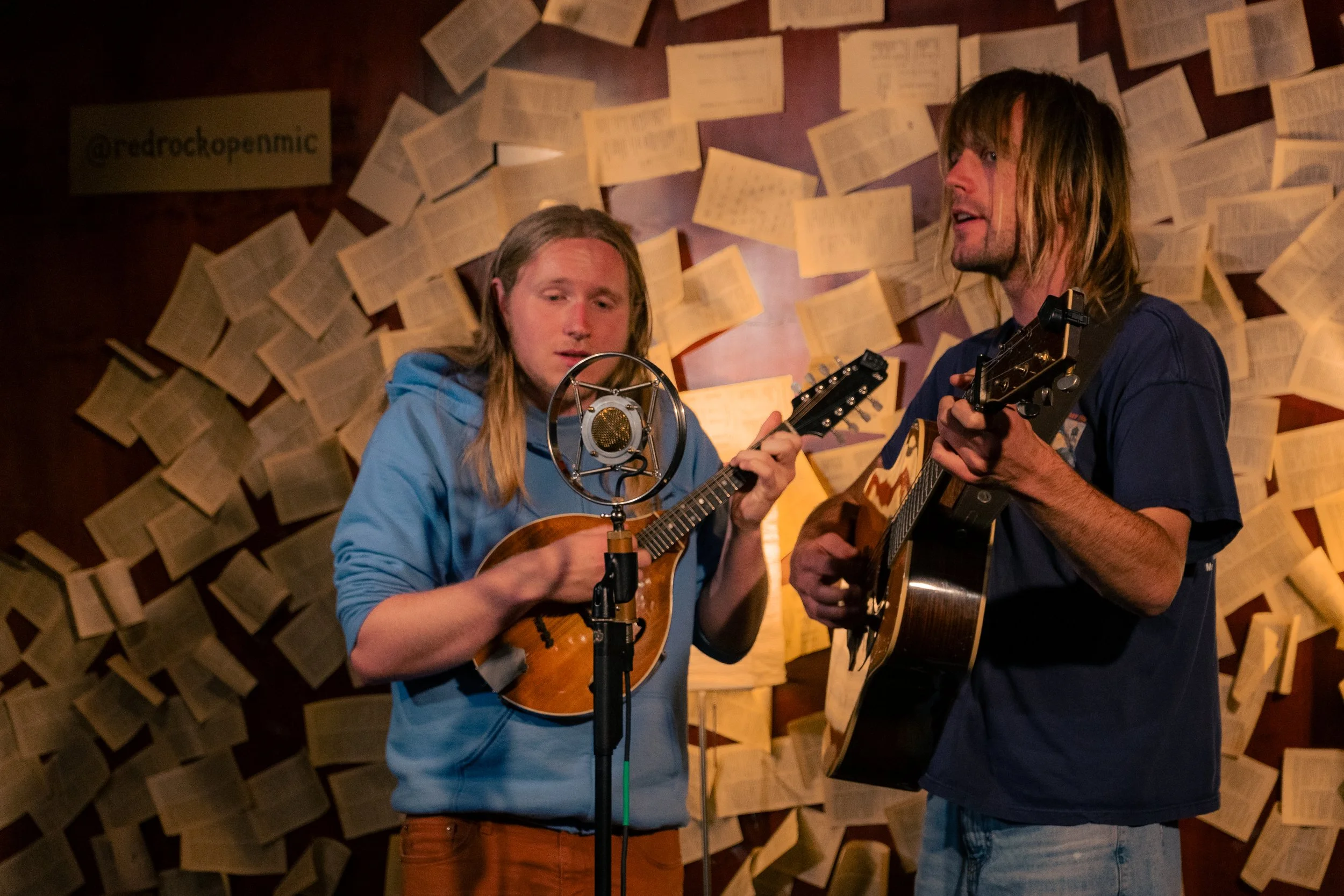 Two men playing guitars and singing in a room with wall decorated with open books.