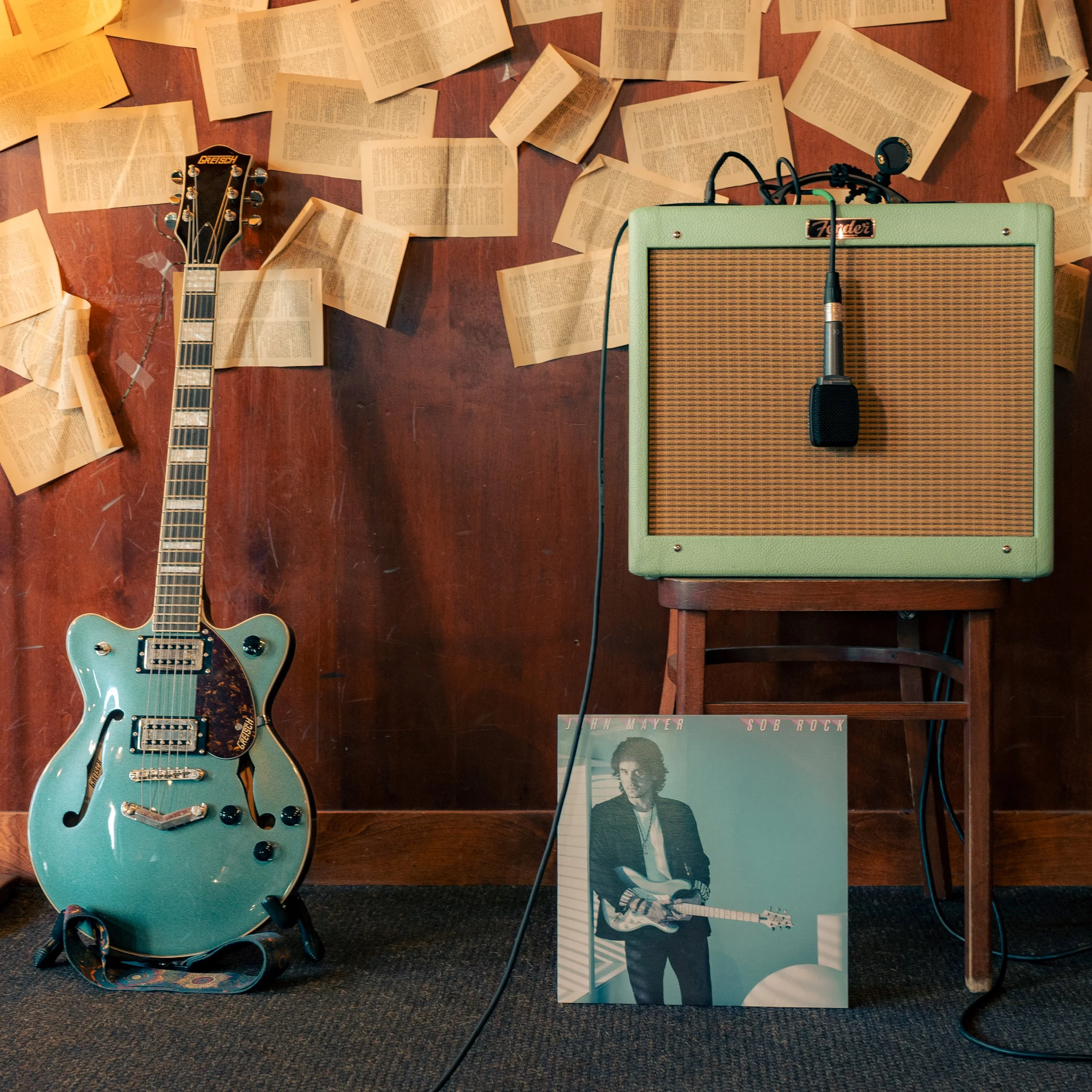 An electric teal guitar leaning against a wooden wall, surrounded by open books, a green Fender amplifier with a microphone attached, and a vinyl album cover of a man holding a guitar.