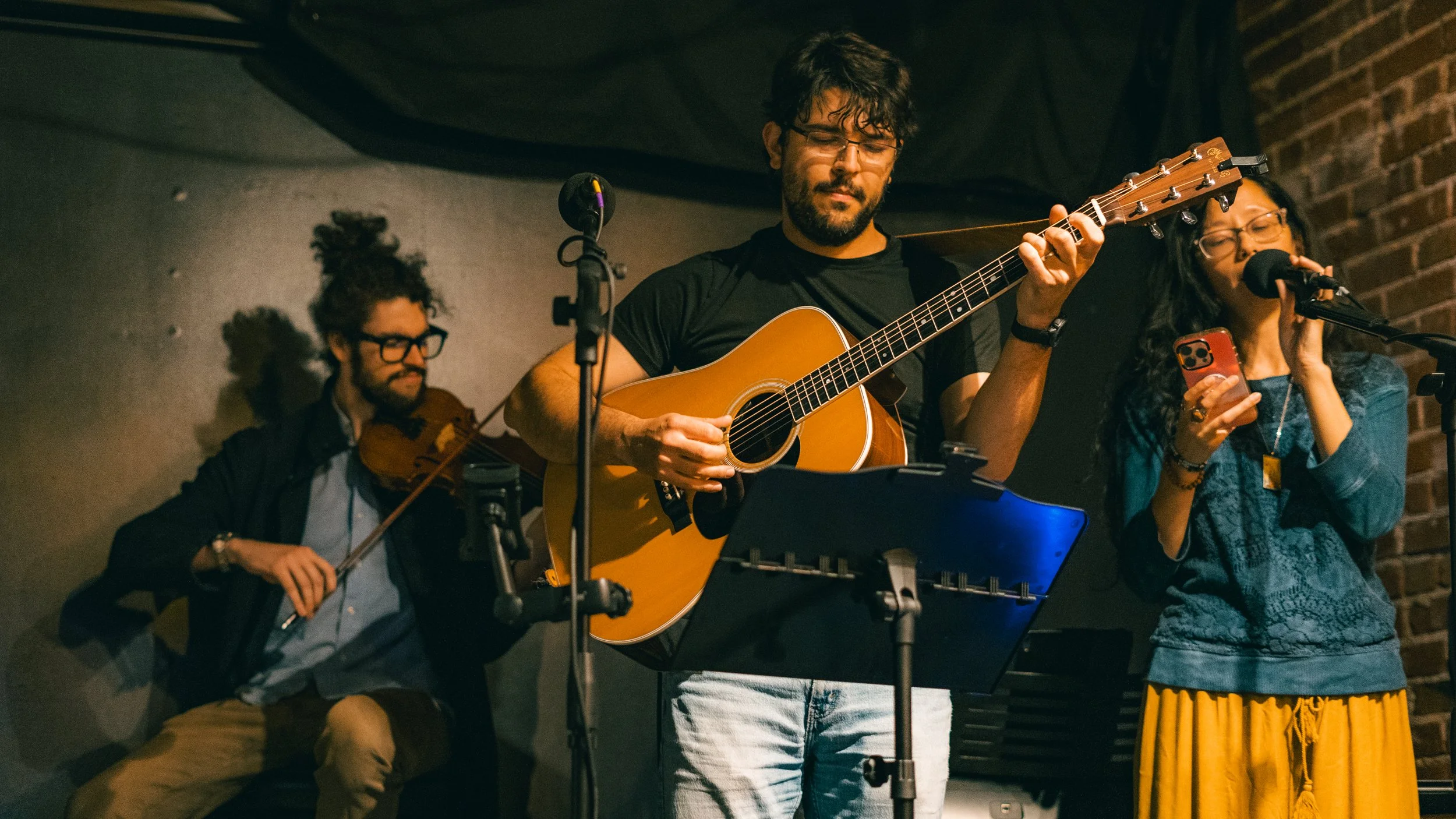Three musicians performing on stage: a man playing an acoustic guitar, a woman singing into a microphone and holding a phone, and a man playing the violin in the background. The setting appears intimate with a brick wall backdrop.