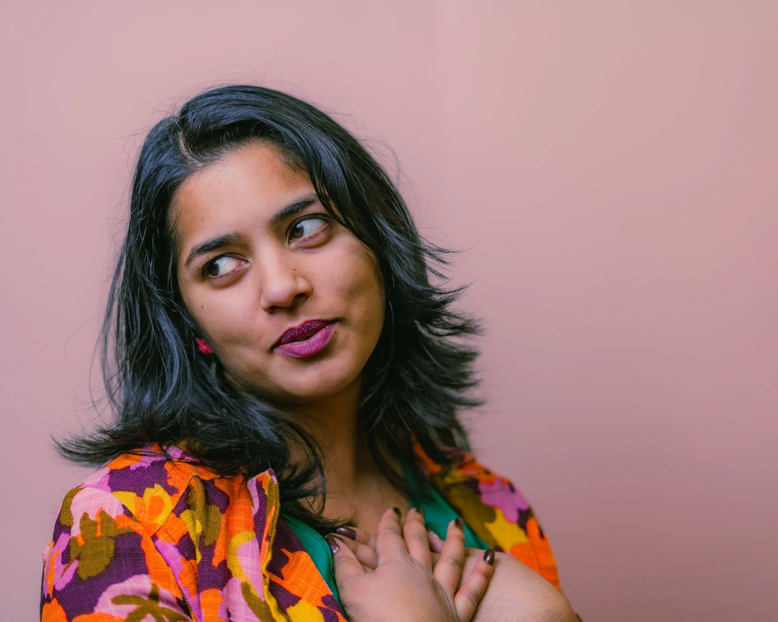 A woman with black hair and dark lipstick is looking to the side, wearing a colorful floral jacket over a green top, with one hand on her chest, against a plain light pink background.