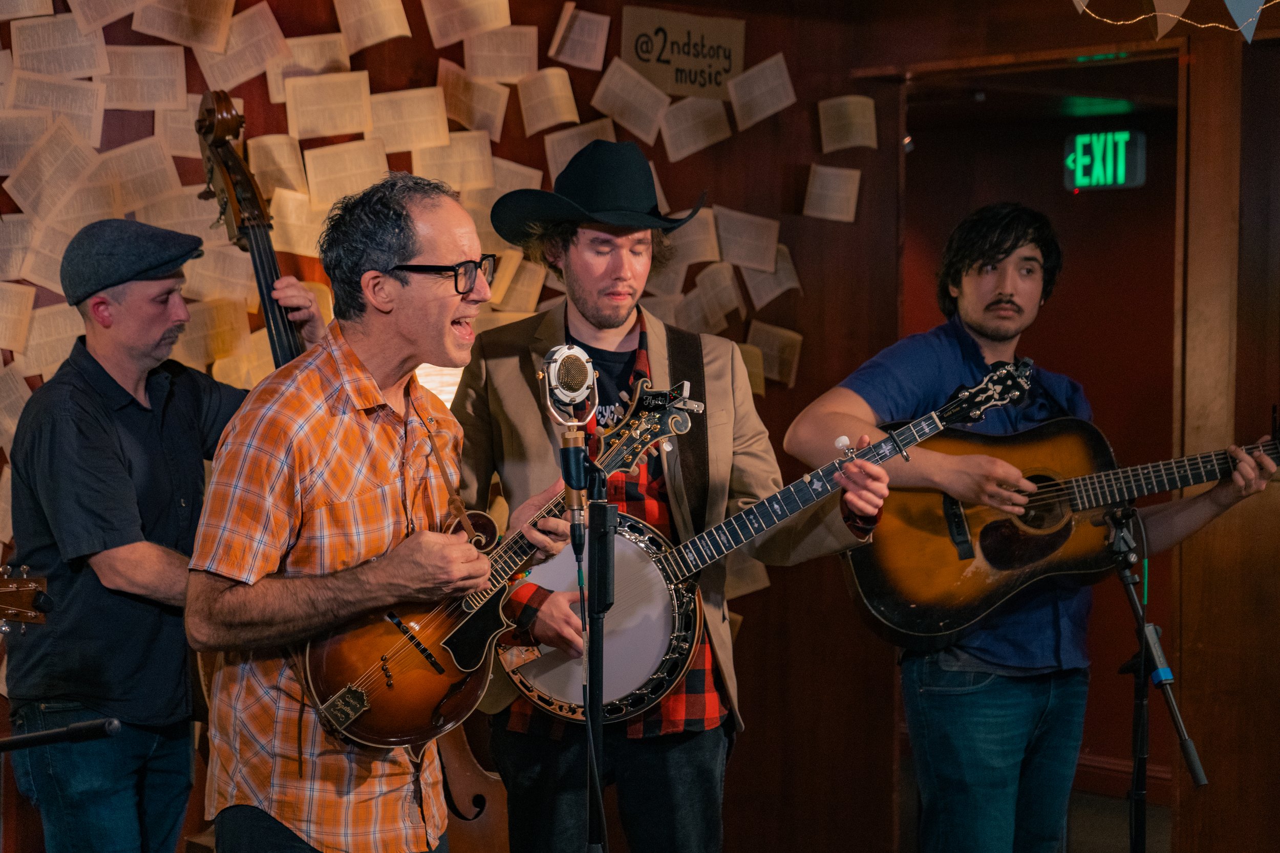Four musicians performing on stage with guitars and a banjo, in front of a wall decorated with open books.