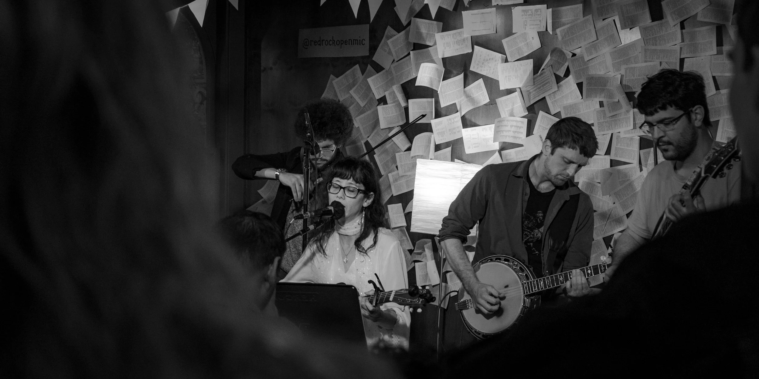 A black and white photo of a musical performance with four musicians. One woman is singing into a microphone and playing a ukulele, while a man next to her plays a banjo. another man is playing a guitar, and a fourth musician is adjusting equipment b