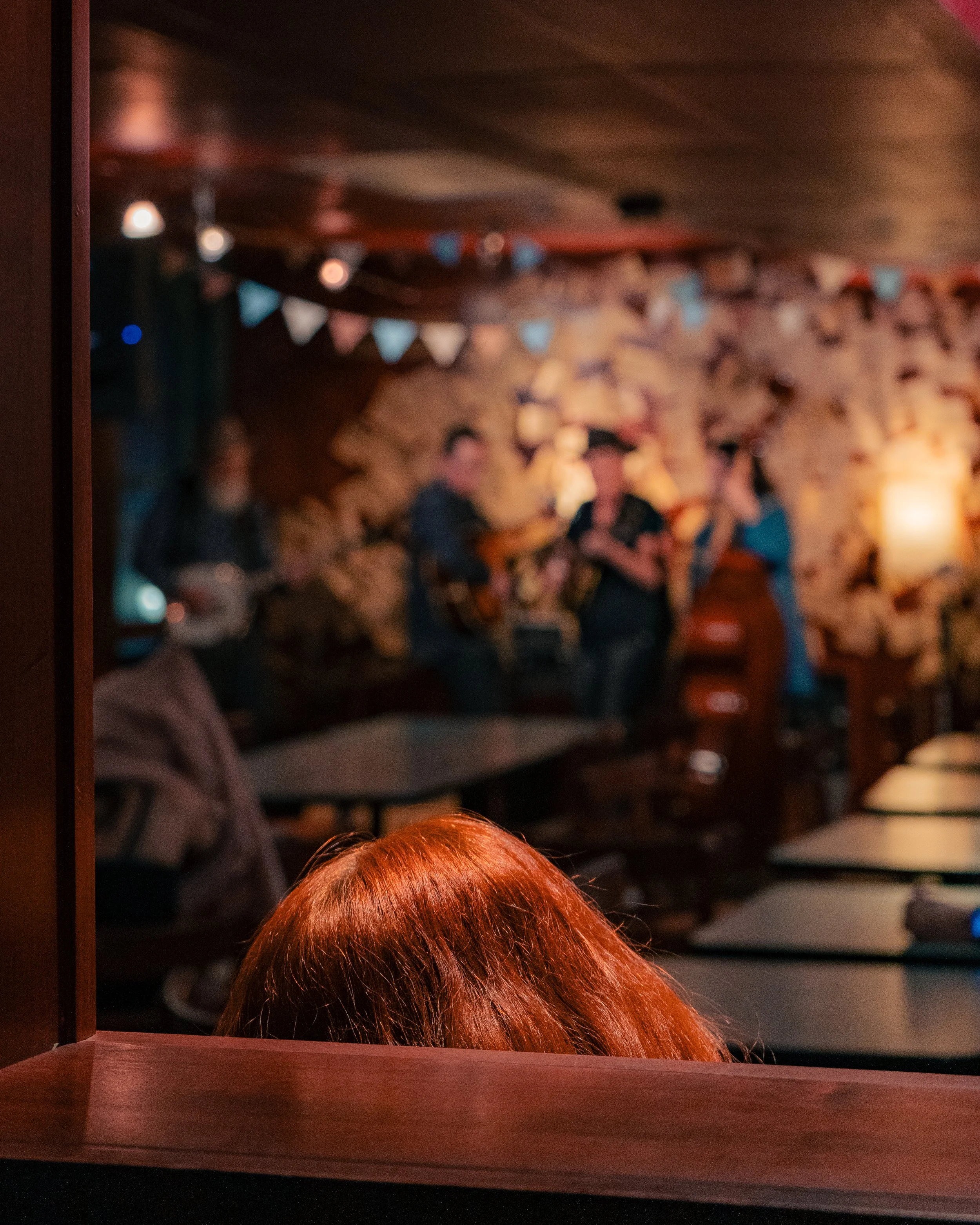 The photo shows the back of a person's head with reddish hair in the foreground, sitting in a dimly lit room with a brick wall and bunting in the background. In the background, there are blurry figures of a band performing, including singers and musi