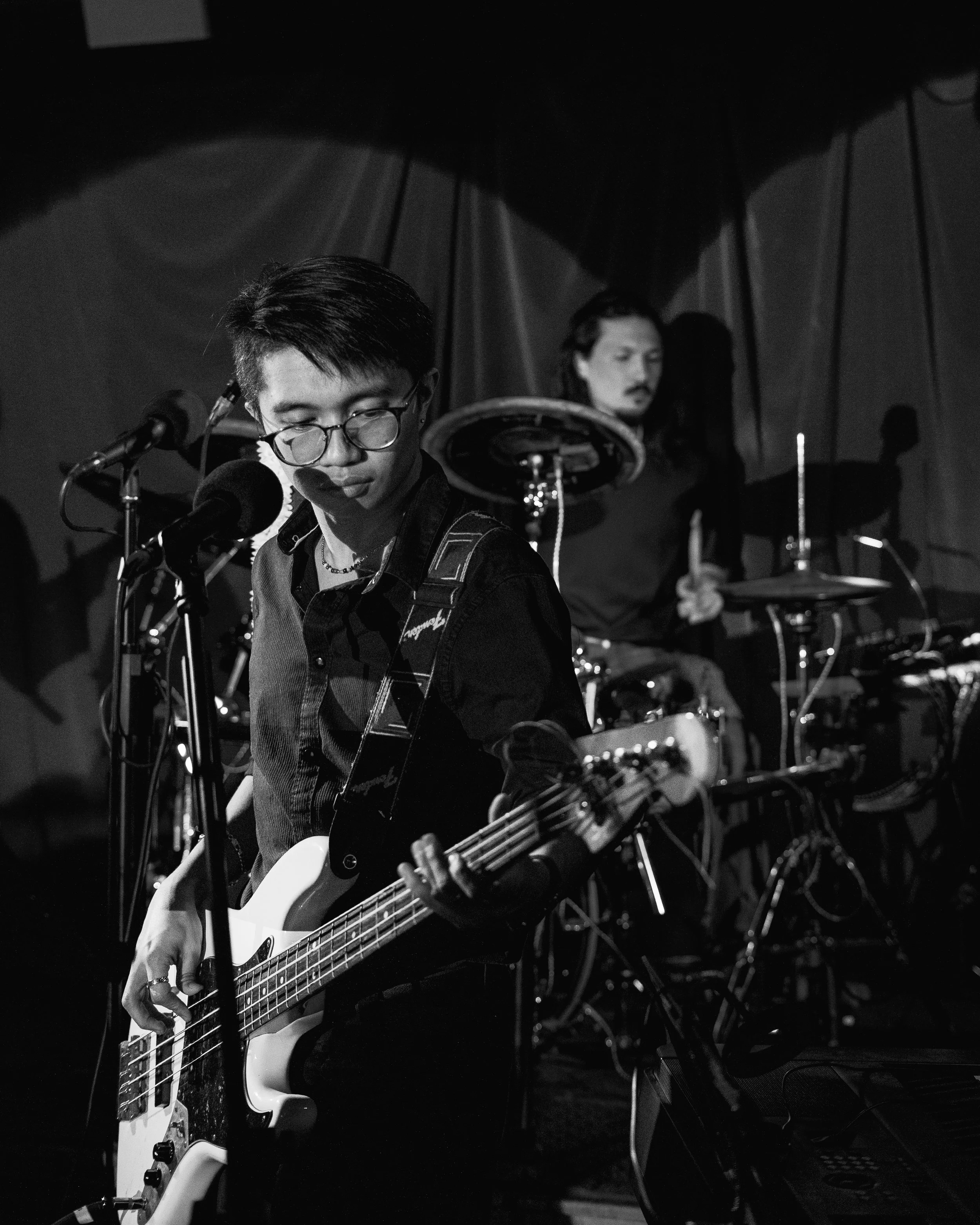 A black-and-white photo of a musician playing a bass guitar and wearing glasses, with a drummer in the background.