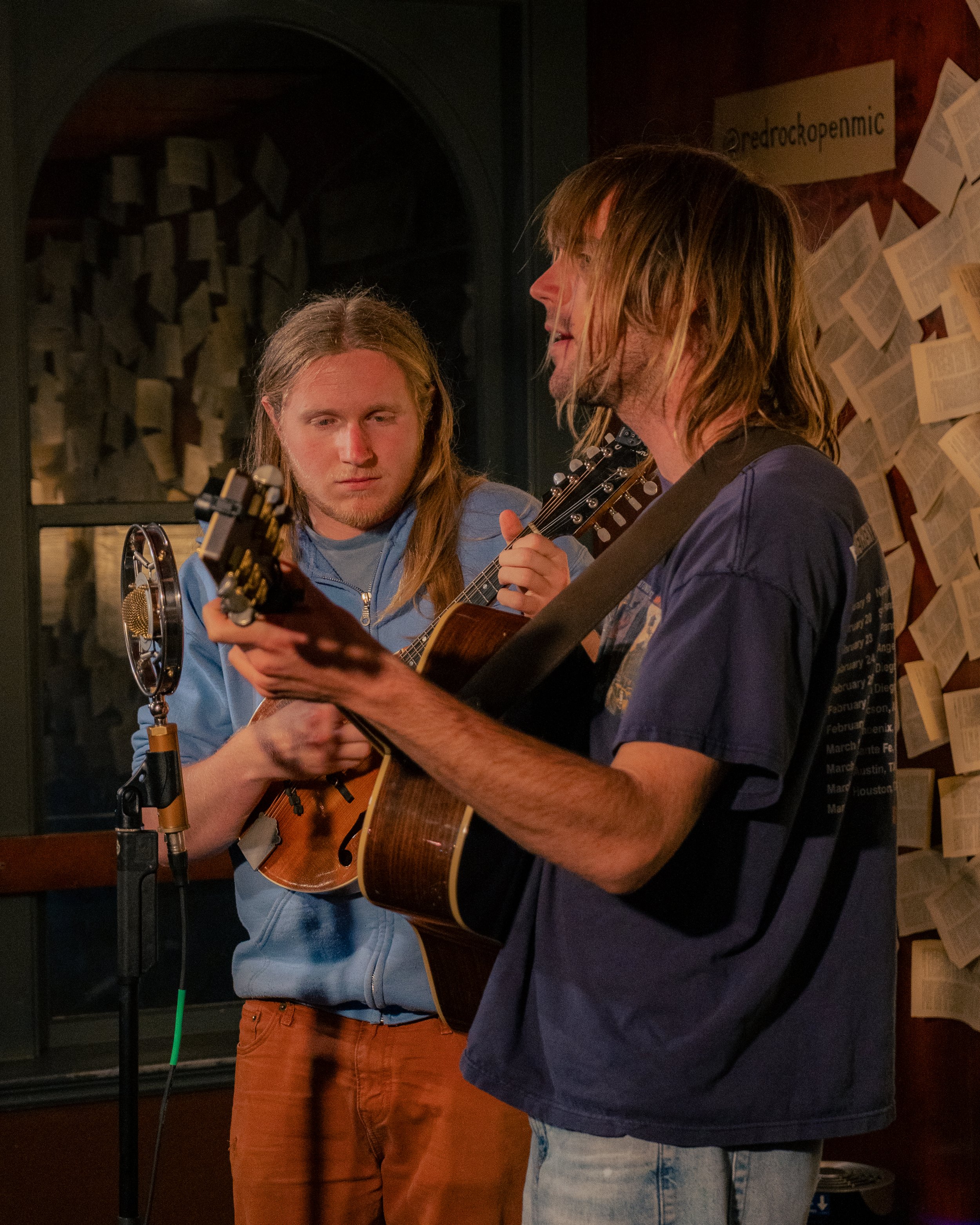 Two young men playing acoustic guitars and singing into a microphone in a dimly lit room with papers on the wall.