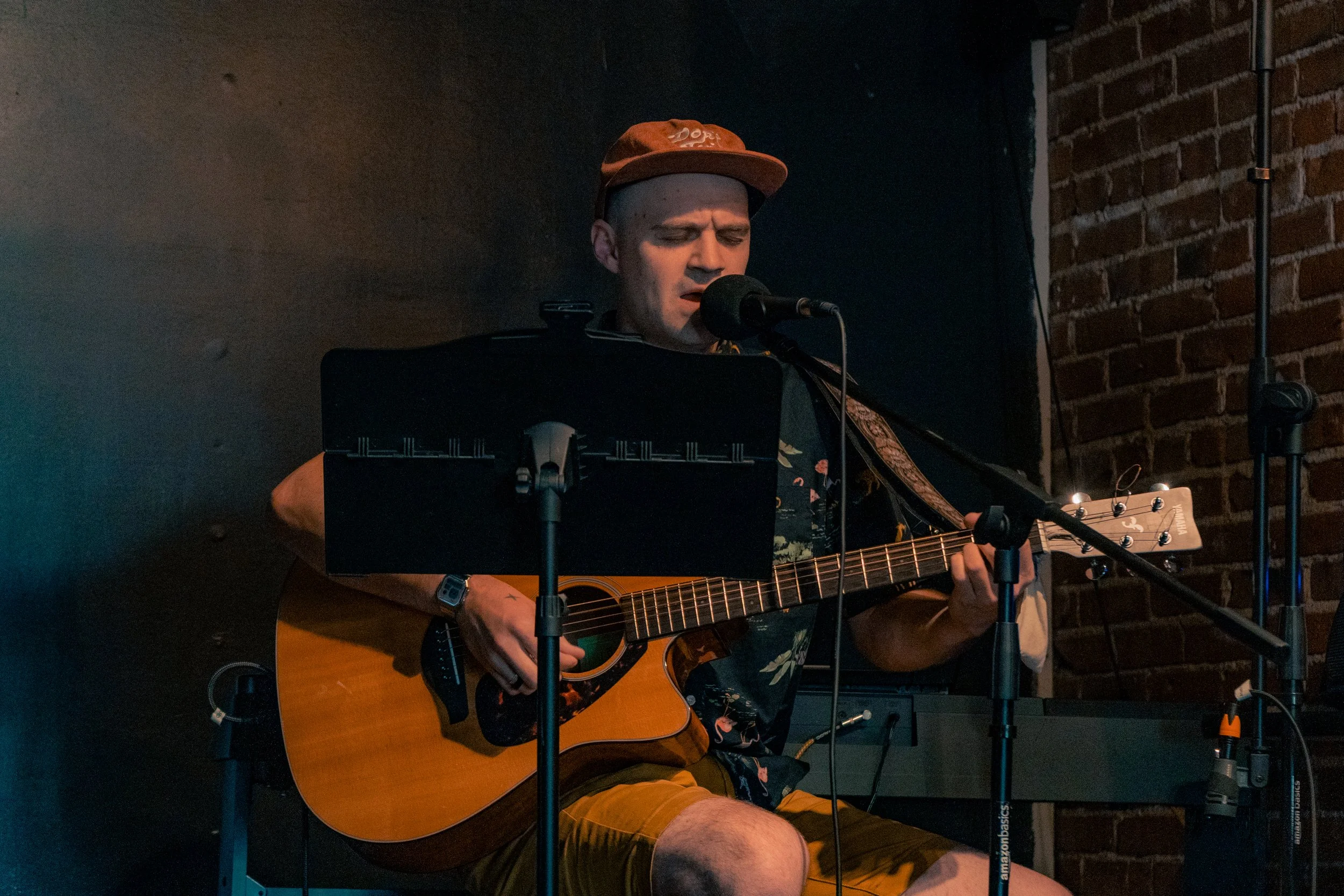 A man wearing a cap and a Hawaiian shirt is playing an acoustic guitar and singing into a microphone on a small stage with brick walls.